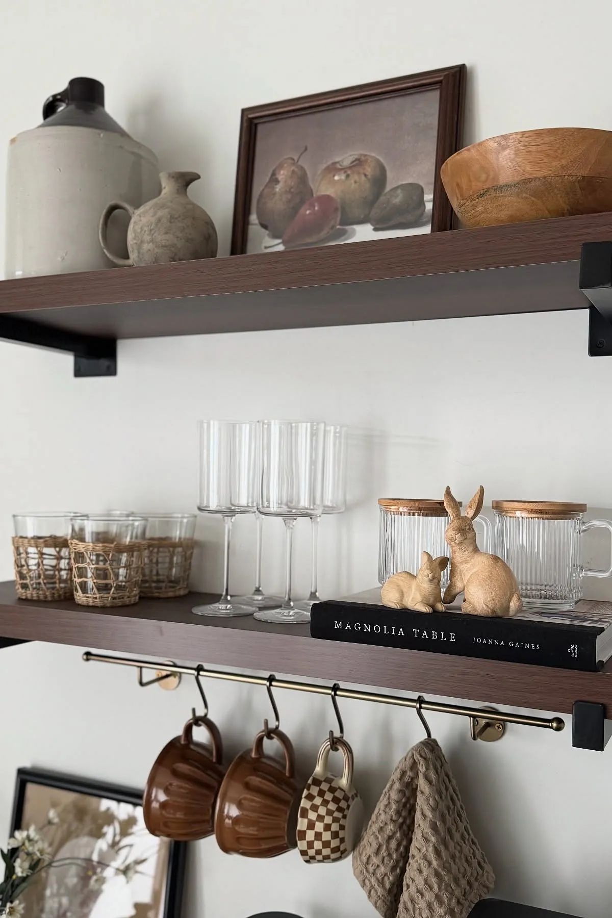 Wooden shelves with ceramic jugs, framed art, glassware, mugs hanging from hooks, and decorative bunnies.