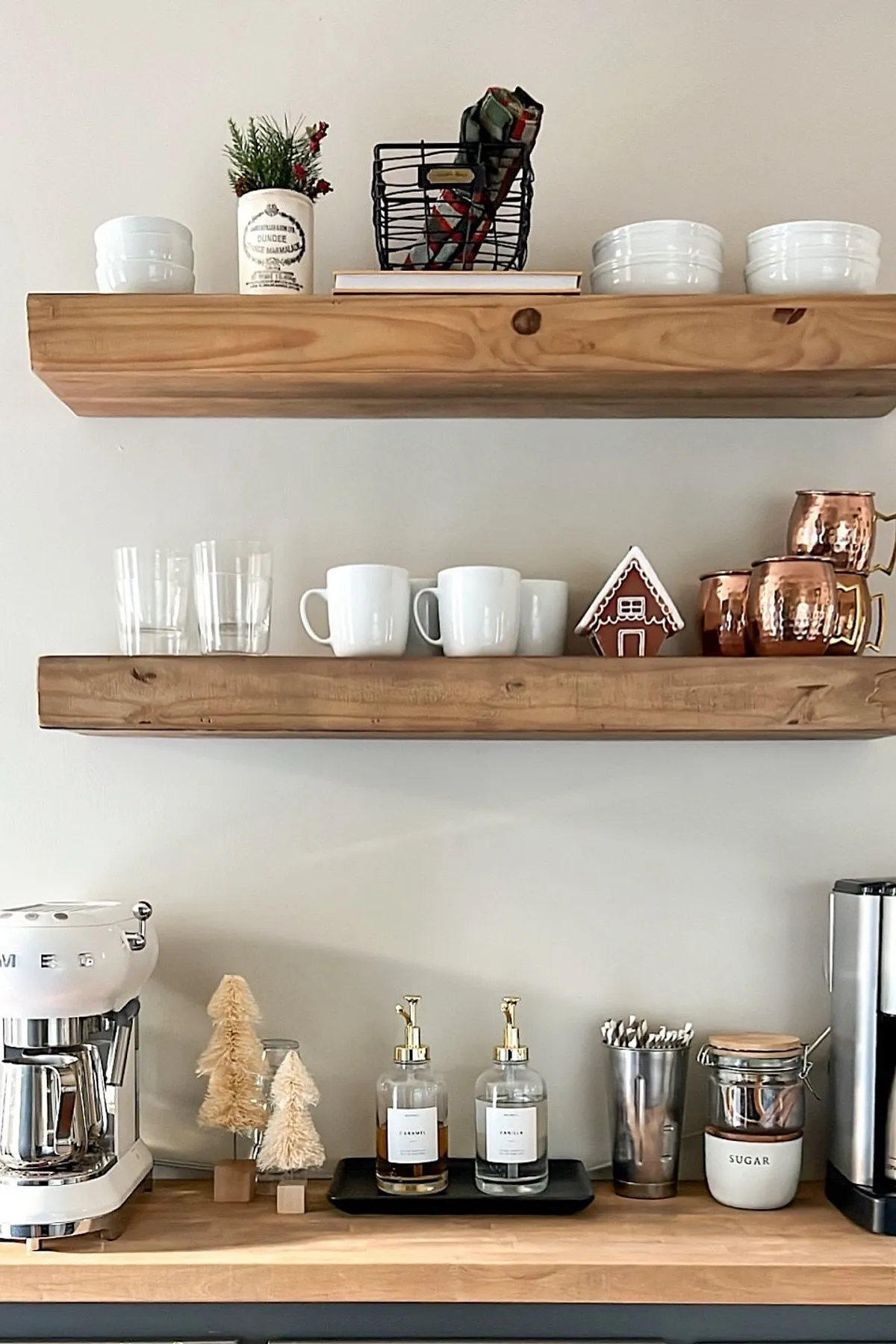 Wooden shelves with mugs, copper cups, syrup bottles, mini trees, and a gingerbread house.