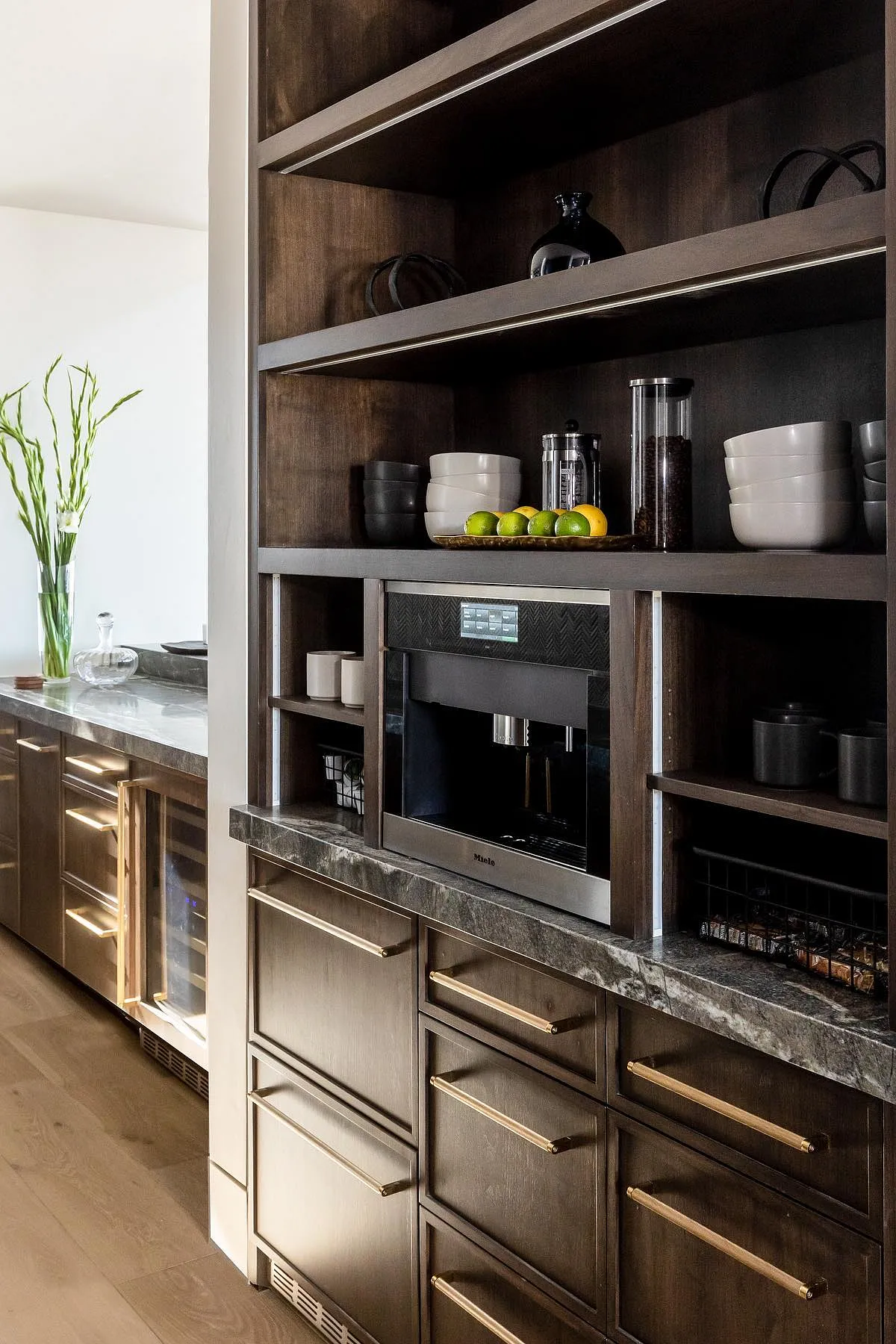Dark wood kitchen coffee station with brass handles, shelves with black and white dishes, and an espresso machine.