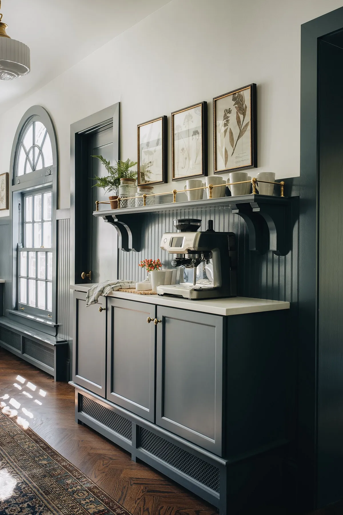 Coffee station with gray cabinets, espresso machine, mugs on a shelf, and framed botanical art above.