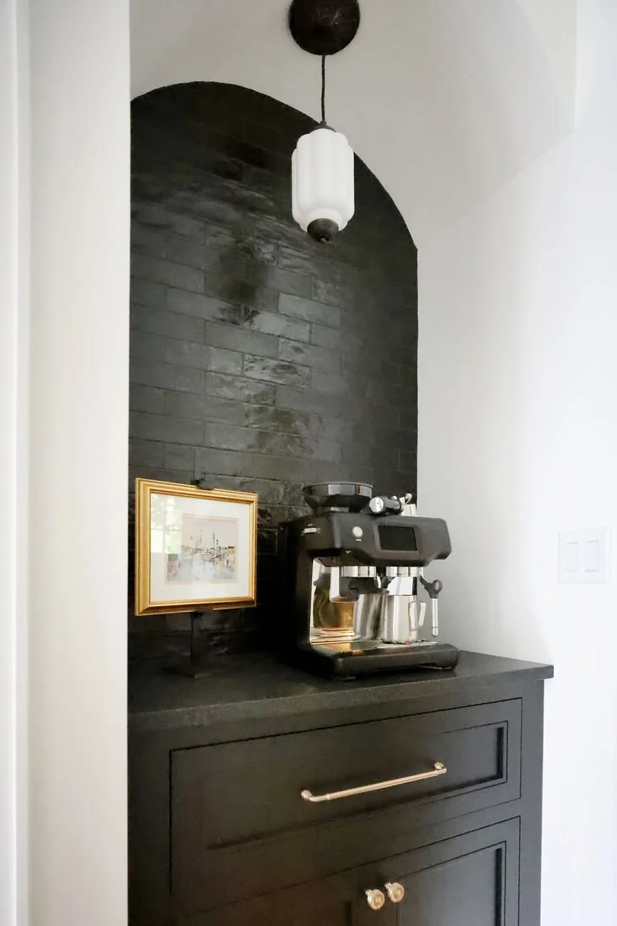 Small coffee station with black tiled wall, espresso machine, framed art, and pendant light.
