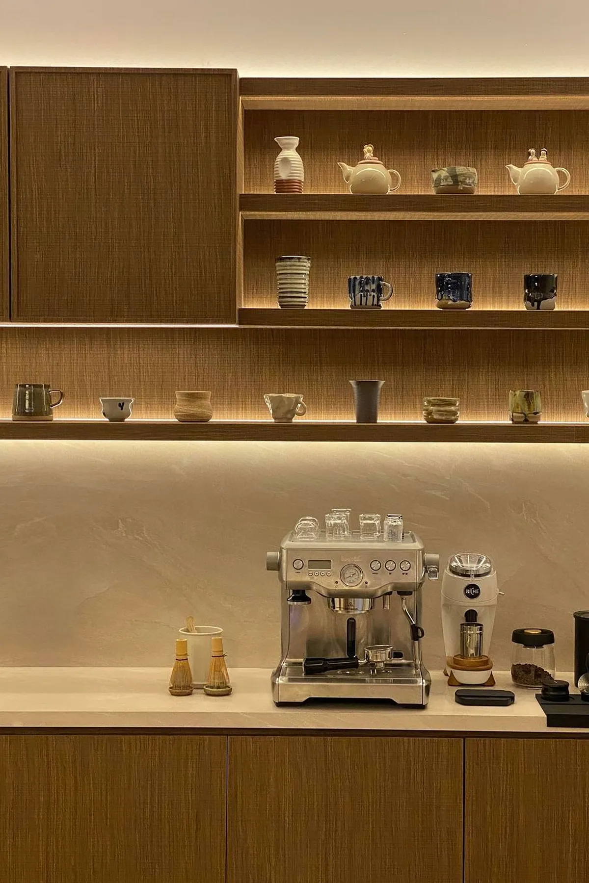 Coffee bar with lit open shelves displaying various ceramic mugs above an espresso machine.