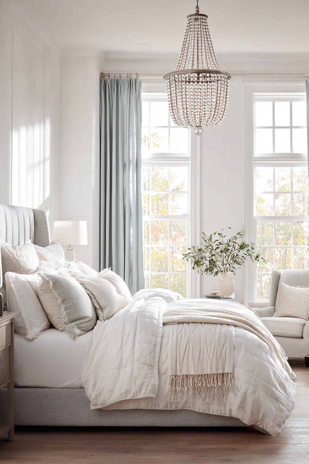 Bedroom with light blue curtains, white bedding, beaded chandelier, and large windows.