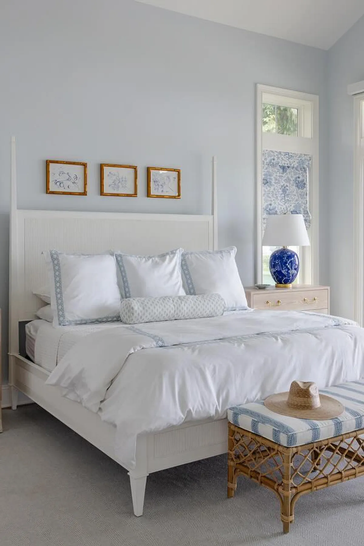 Bedroom with white bed, blue and white lamp, rattan bench, and framed artwork above the headboard.