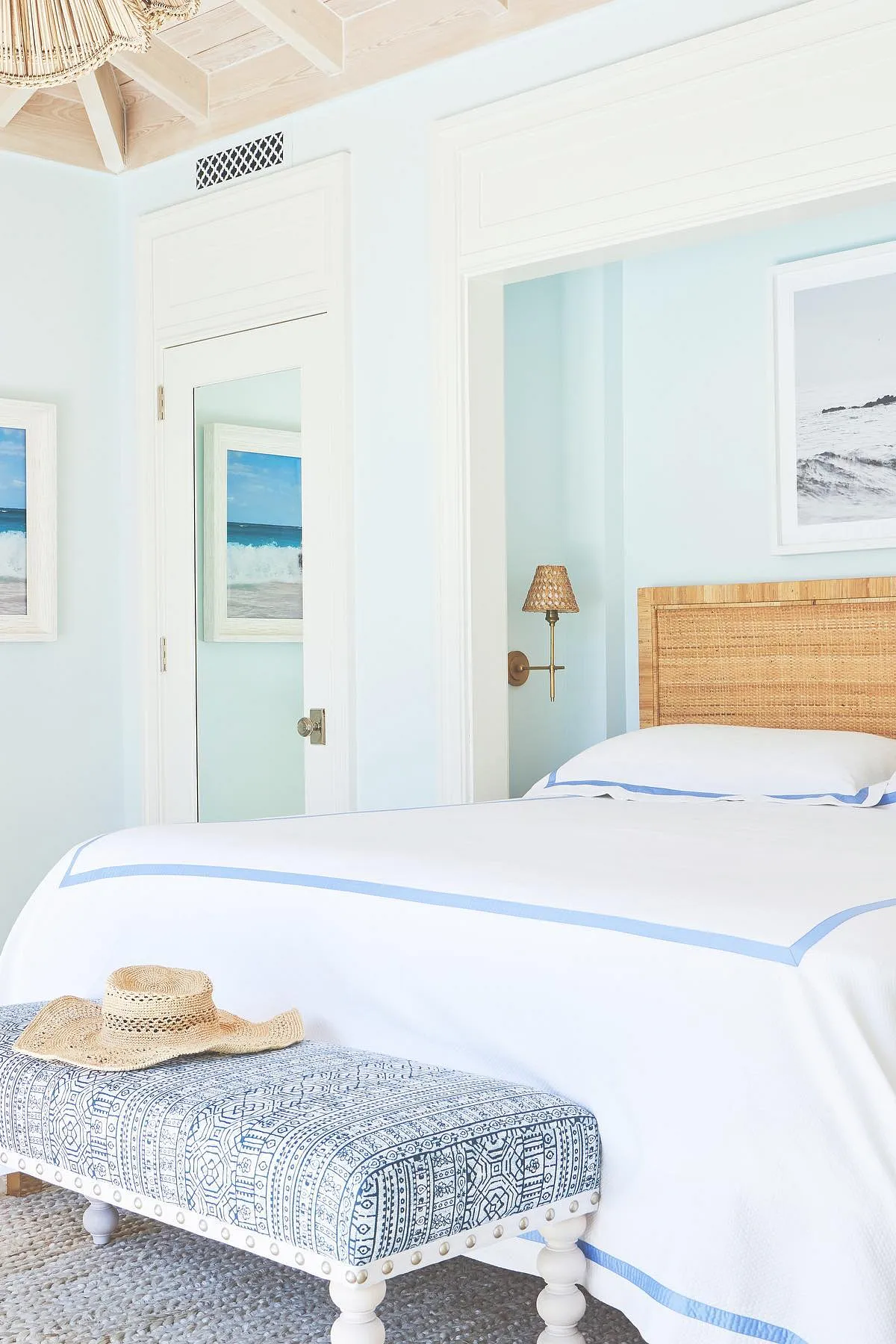 Bedroom with light blue walls, rattan headboard, white bedding, patterned bench, and ocean-themed artwork.