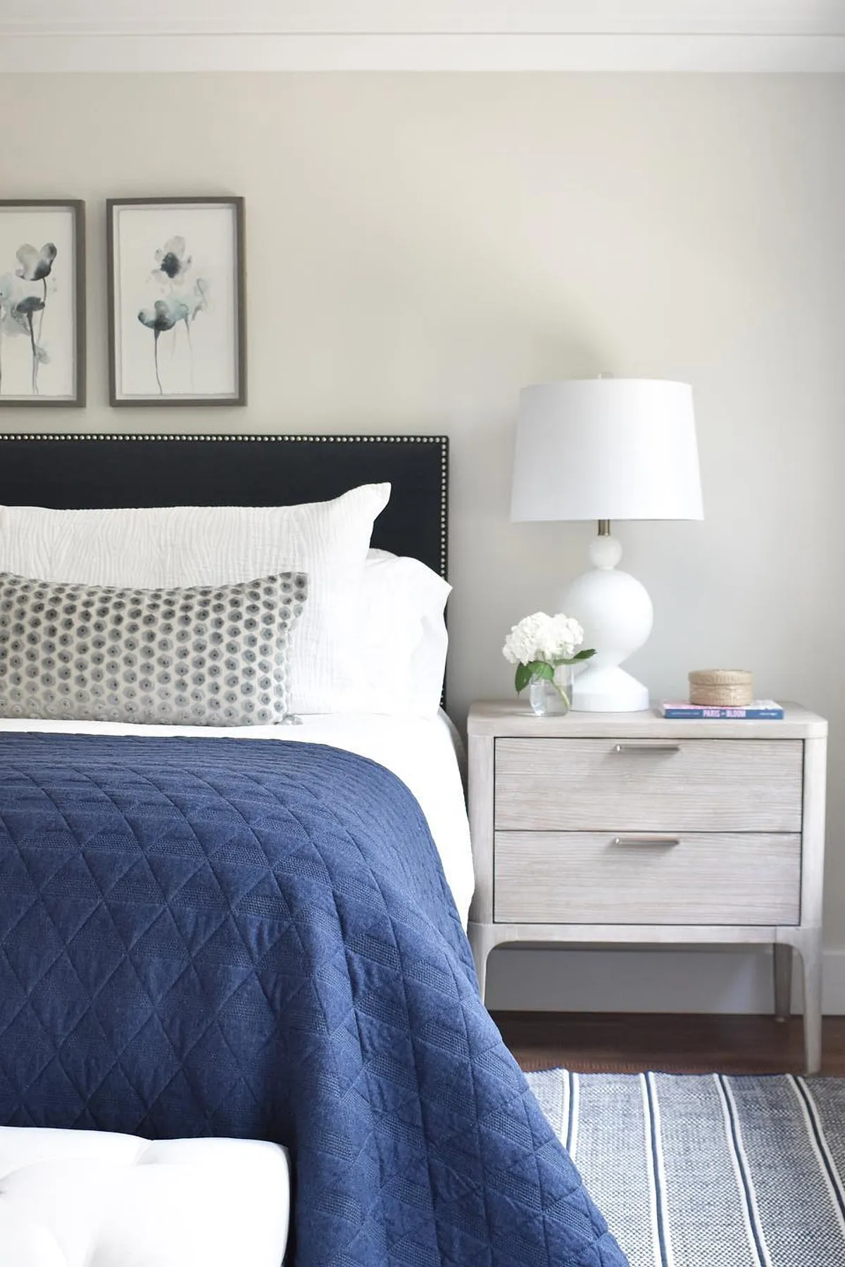 Bedroom featuring navy quilt, white bedding, floral artwork, upholstered headboard, and a white lamp on a nightstand.