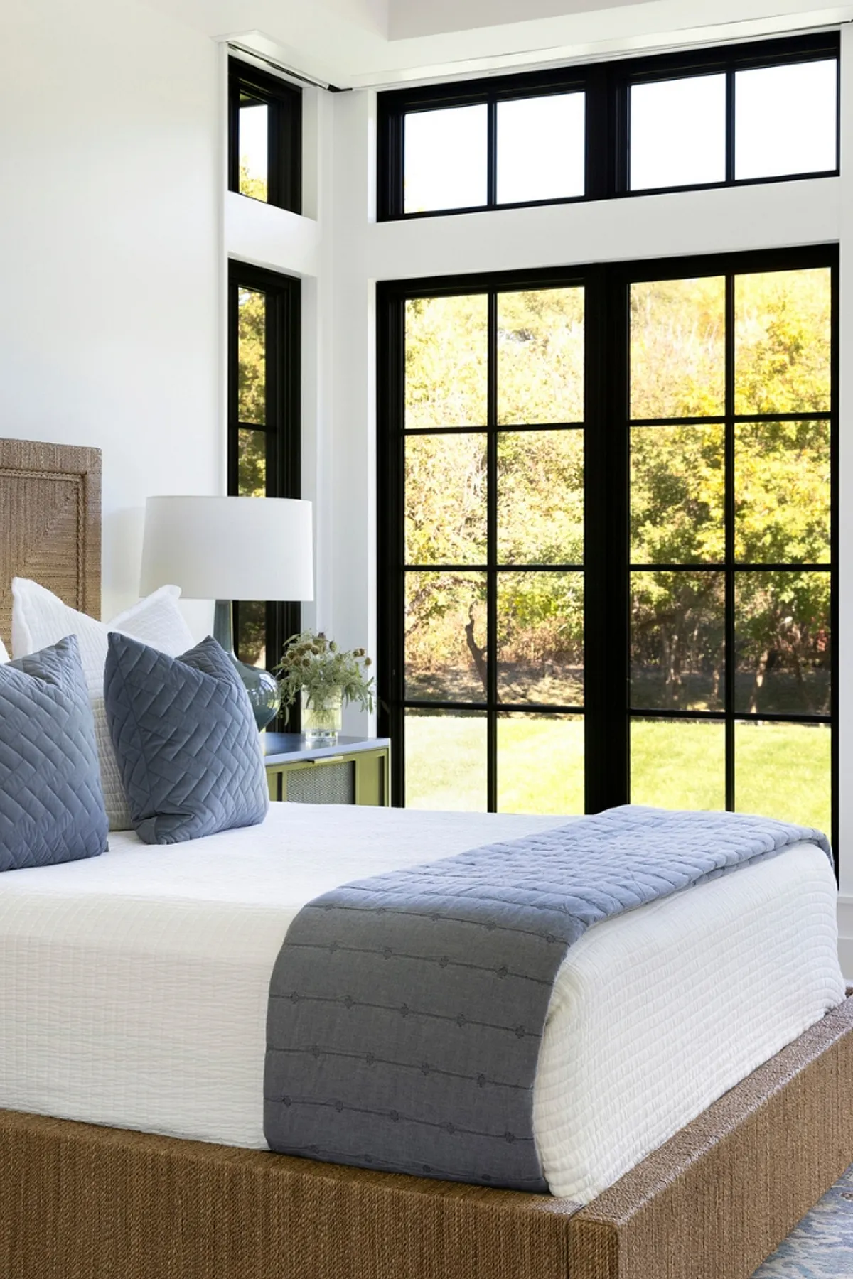 Bedroom with large windows, white bedding, muted blue pillows and bed runner, wicker headboard.