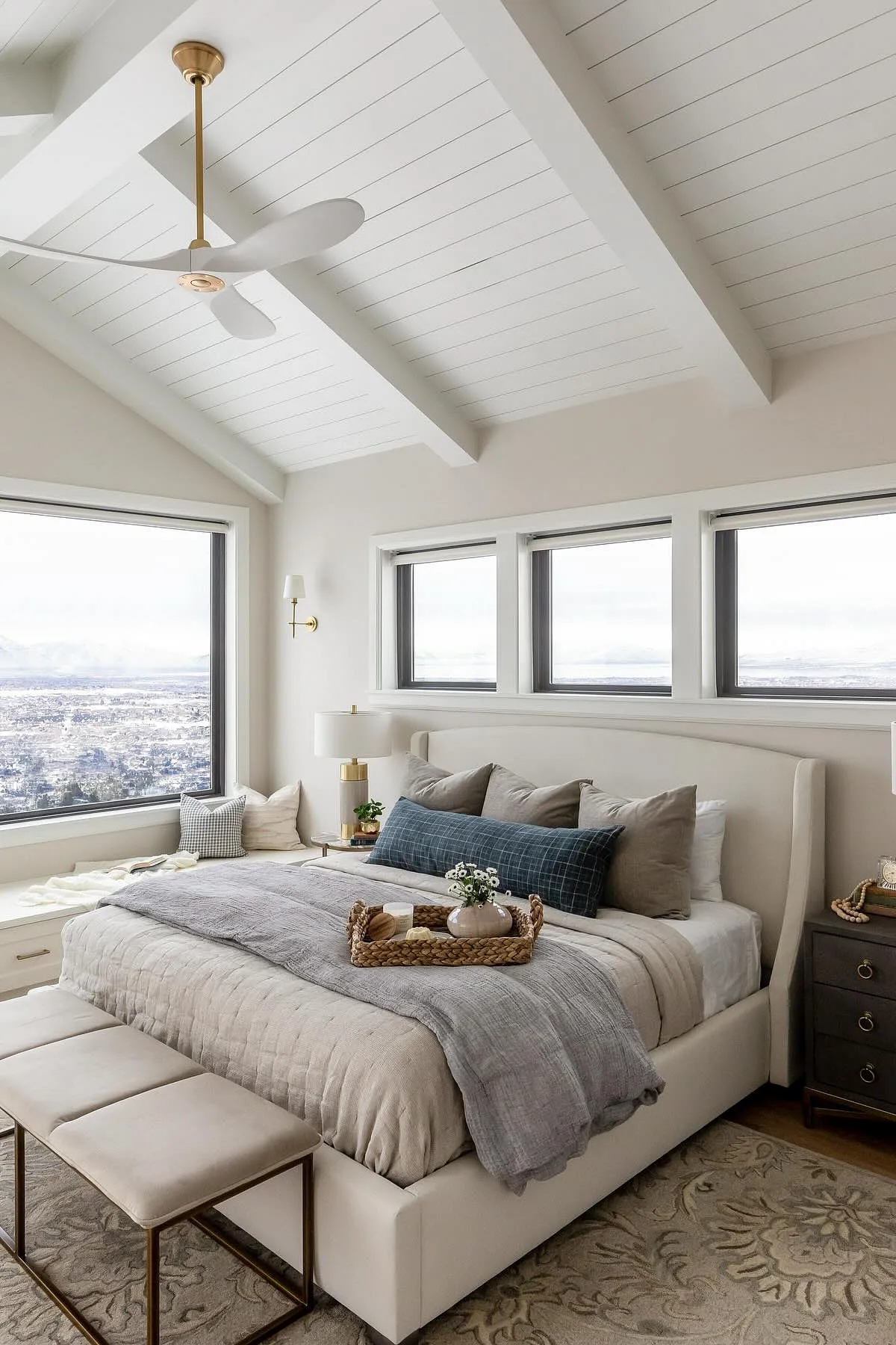 Neutral bedroom with large windows, white bedding, blue accent pillows, and window bench.