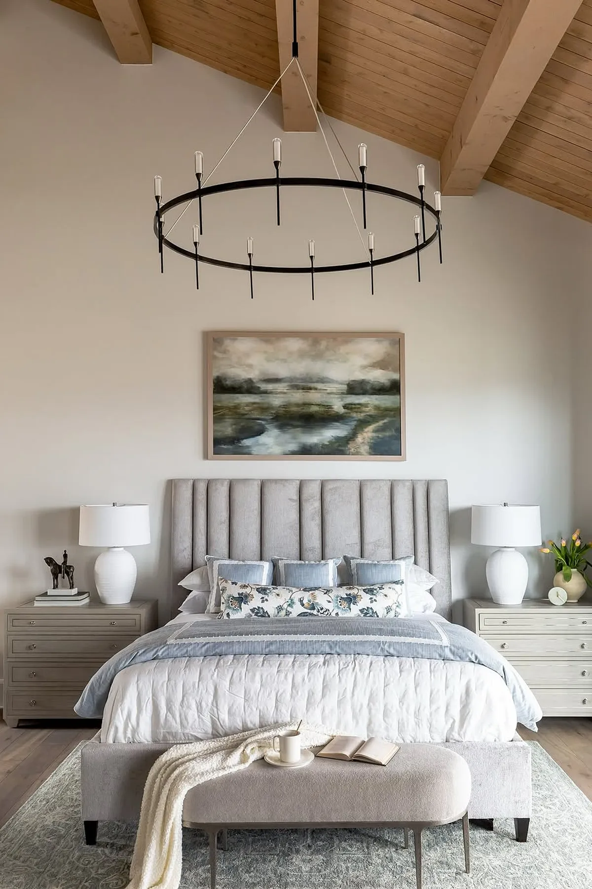 Bedroom with light wood ceiling, large chandelier, abstract art, light blue accents on white bedding.