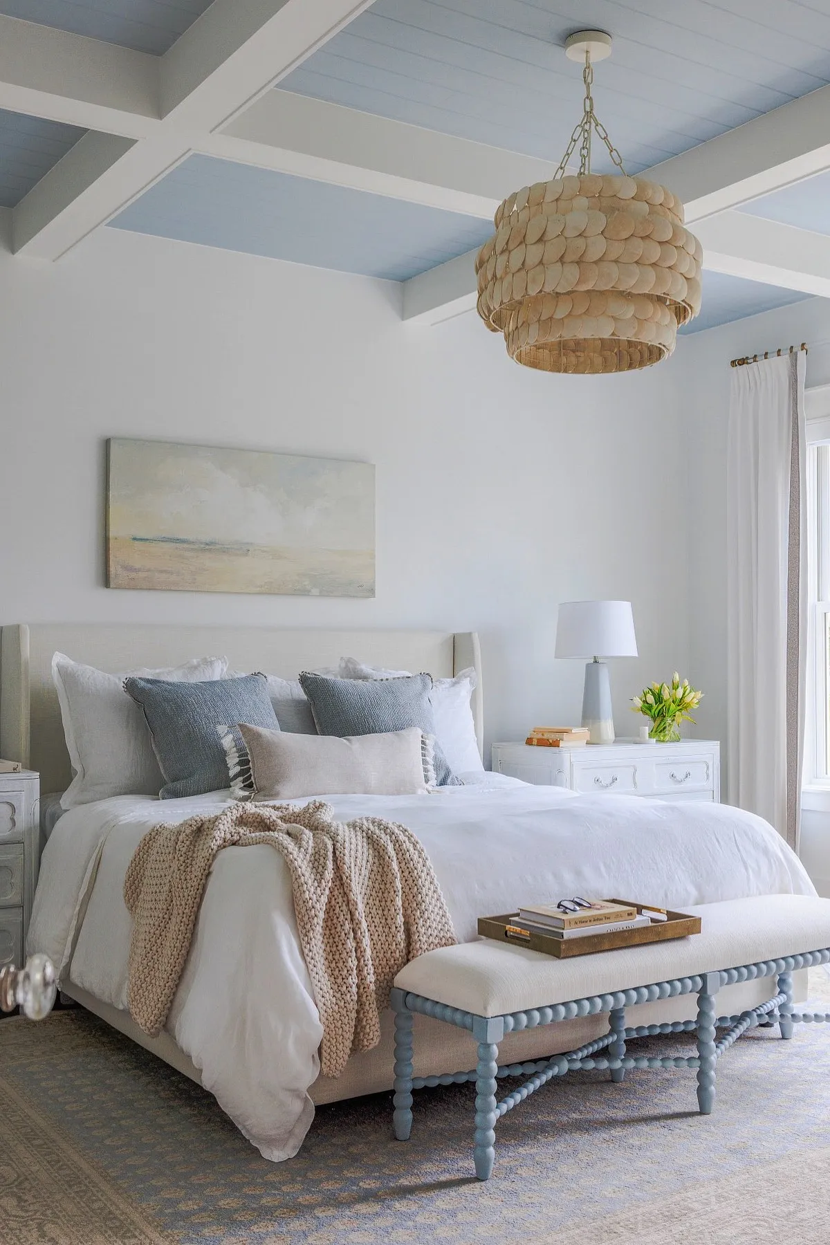 Bedroom with light blue ceiling, white bedding, textured chandelier, and muted artwork above the bed.