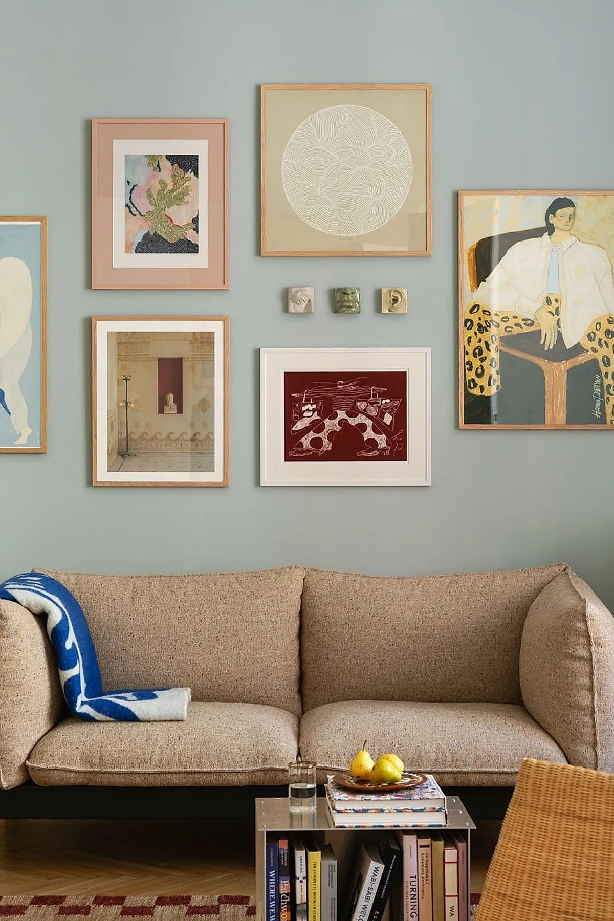 Pastel and earthy toned artwork on a blue wall above a beige sofa with books on a side table.