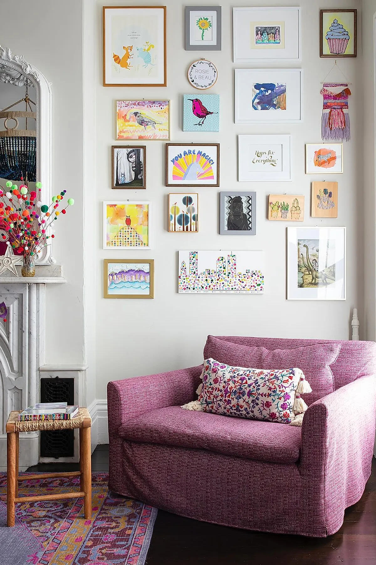 Living room with a vibrant gallery wall of colorful framed art above a magenta chair, next to a decorative fireplace.