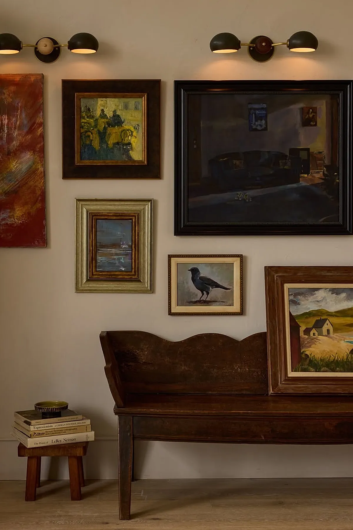 Gallery wall with vintage paintings in various frames above a rustic wooden bench, accented by warm wall lights.
