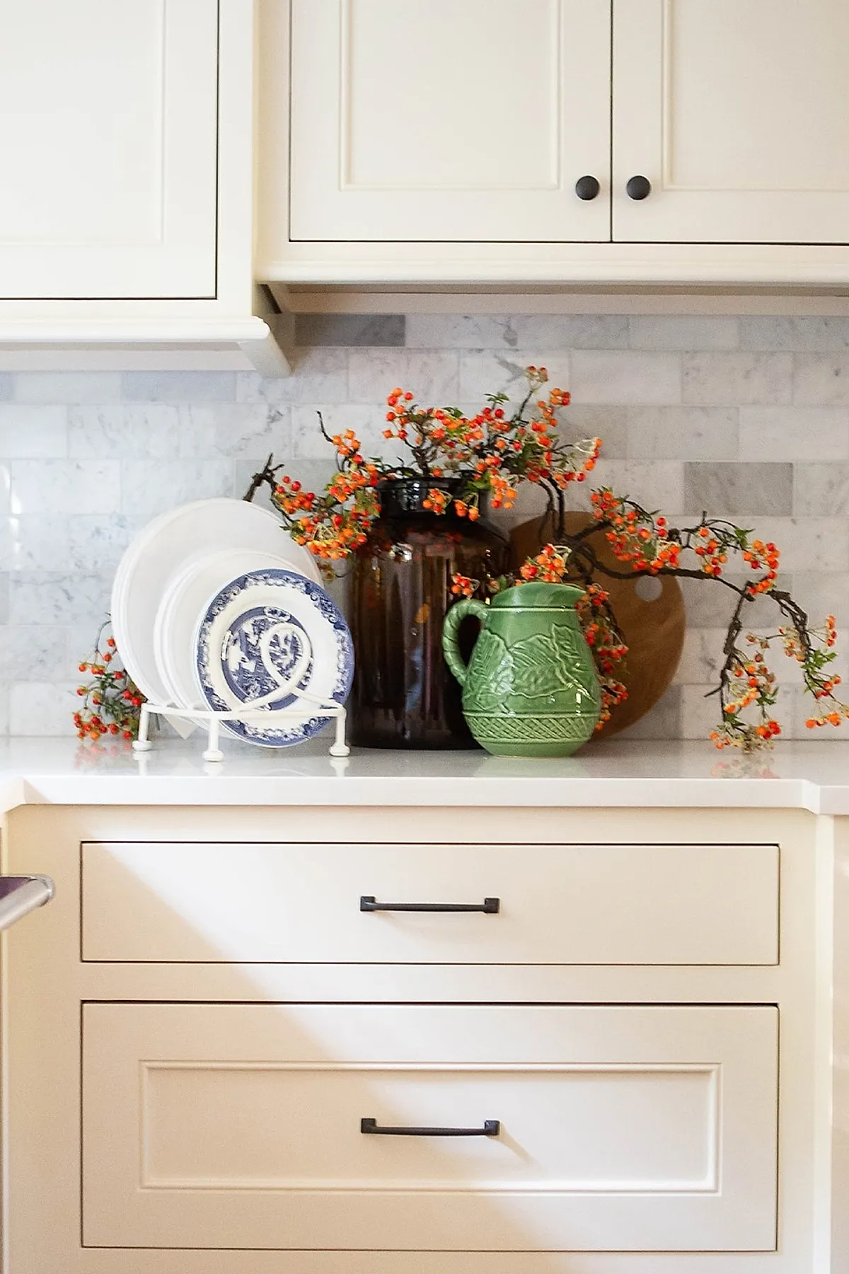 Kitchen countertop with white and blue patterned vintage plates in a white plate rack, a green ceramic pitcher, and orange berry branches spilling from an amber glass vase against cream cabinets and marble tile backsplash.