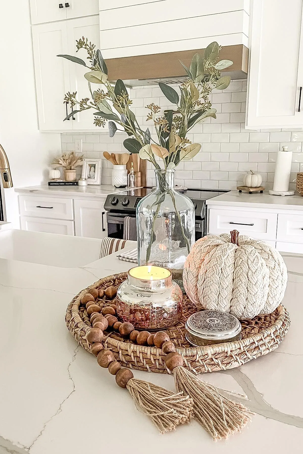 Neutral-toned fall kitchen countertop decor featuring a woven tray holding a cream knitted pumpkin, wooden bead garland with tassels, clear glass vase with green foliage, and metallic candle in a bright white kitchen.