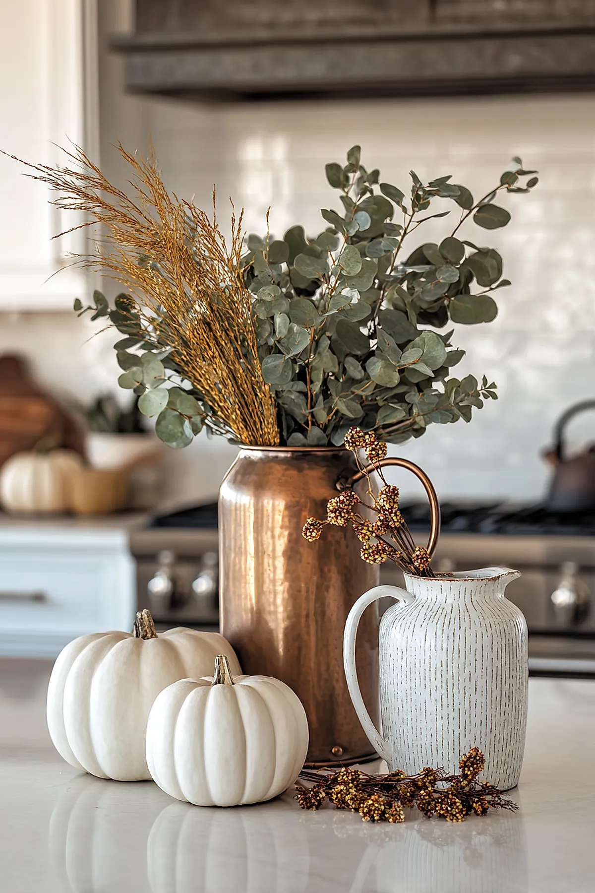 Copper pitcher holding eucalyptus branches alongside two white pumpkins on a light-colored kitchen countertop with blurred stove in the background.