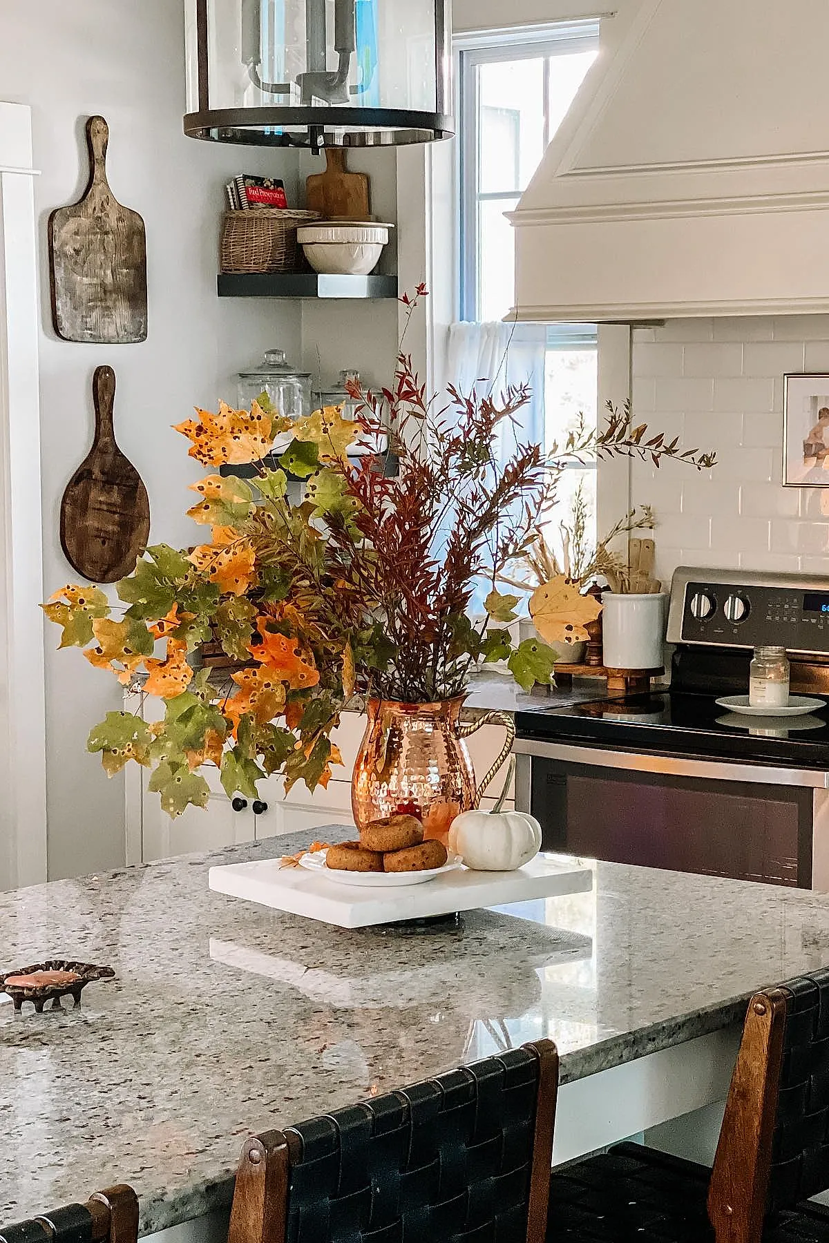 Kitchen island with granite countertop featuring hammered copper pitcher filled with autumn leaves in orange, yellow, and green hues, next to a small white pumpkin and plate of cookies.