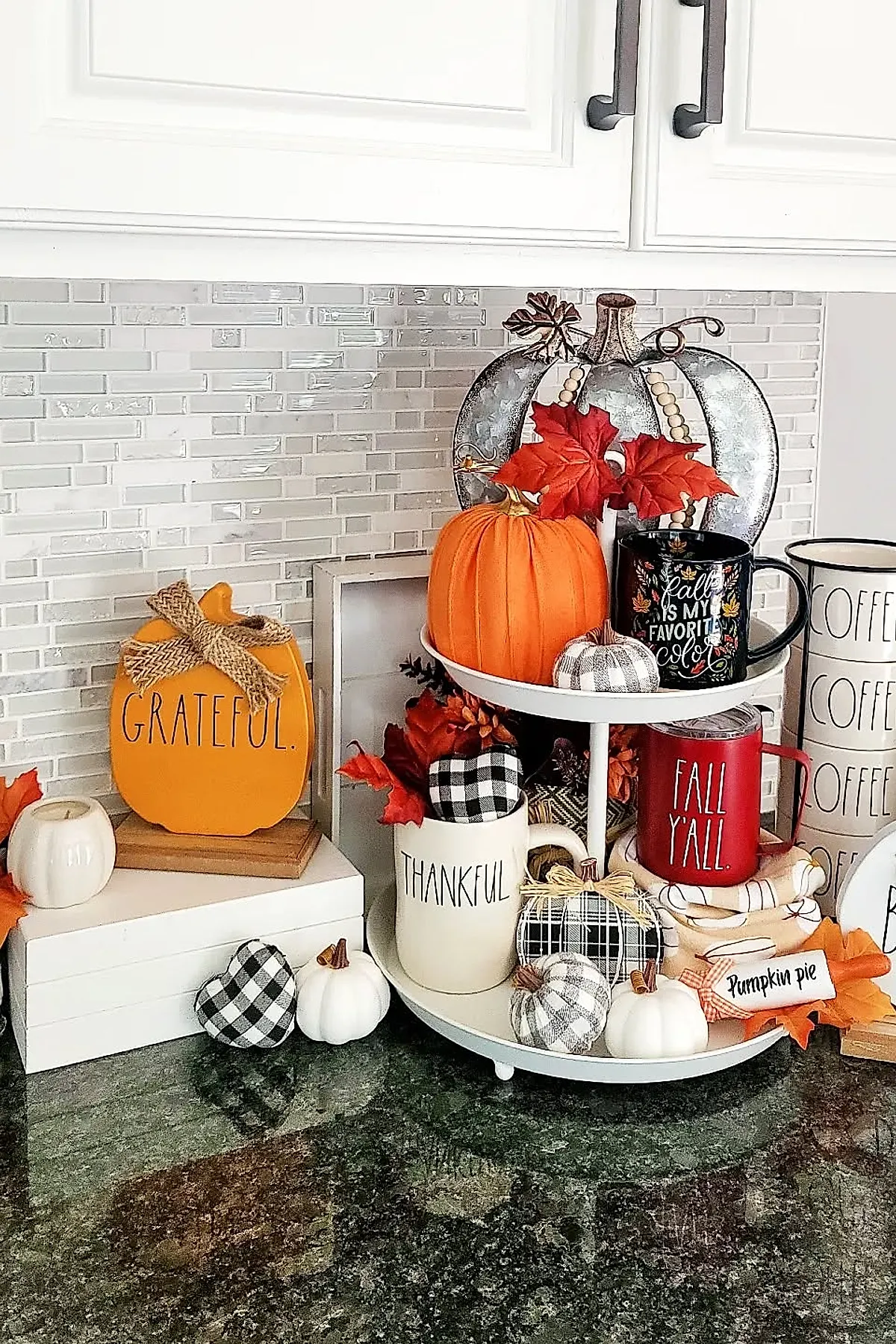 Two-tiered white tray on kitchen counter decorated with orange and white pumpkins, mugs labeled thankful and fall y'all, black-and-white buffalo plaid fabric hearts, faux red leaves, and a small pumpkin pie sign.