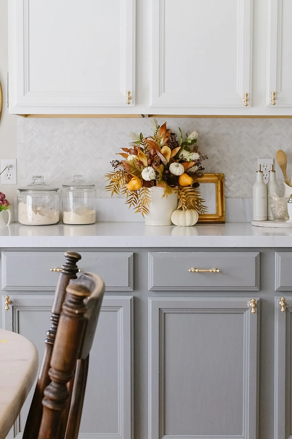 White ceramic vase filled with autumn foliage including golden leaves, pinecones, and muted orange flowers next to small white pumpkins on a gray kitchen countertop with white cabinets.