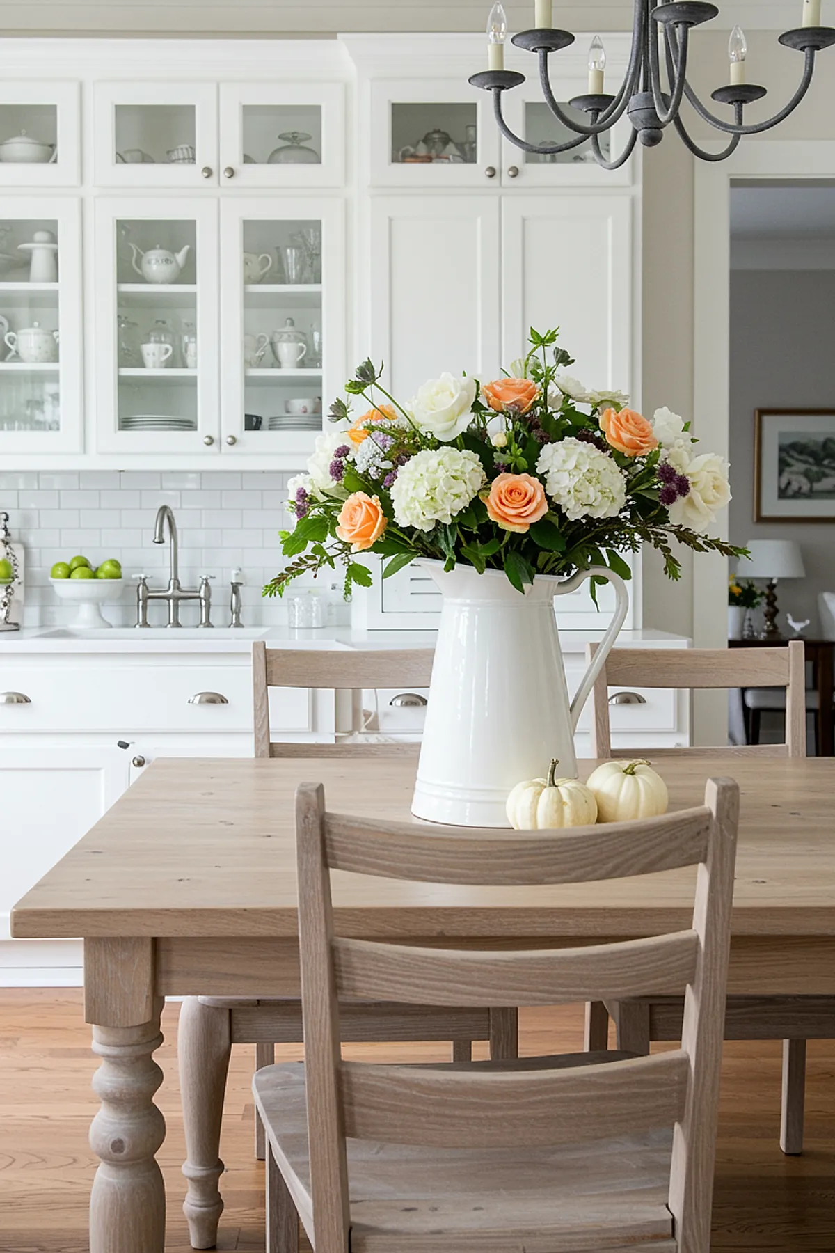 Wooden kitchen table with a white ceramic pitcher filled with cream hydrangeas, peach roses, green foliage, and purple accents. Two small white pumpkins sit next to the pitcher. White cabinets and glass-front cupboards are visible in the background.