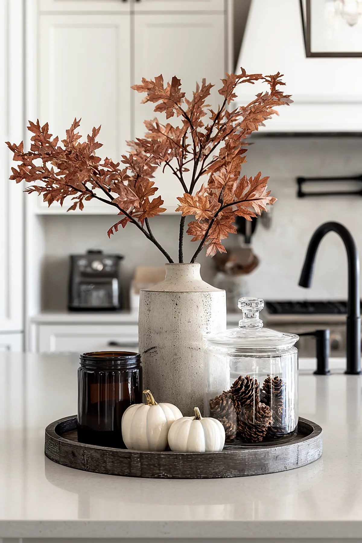 Kitchen countertop featuring a dark wooden tray holding a textured beige ceramic vase with dried brown oak leaves, an amber glass candle holder, two small white pumpkins, and a clear glass jar filled with pinecones.