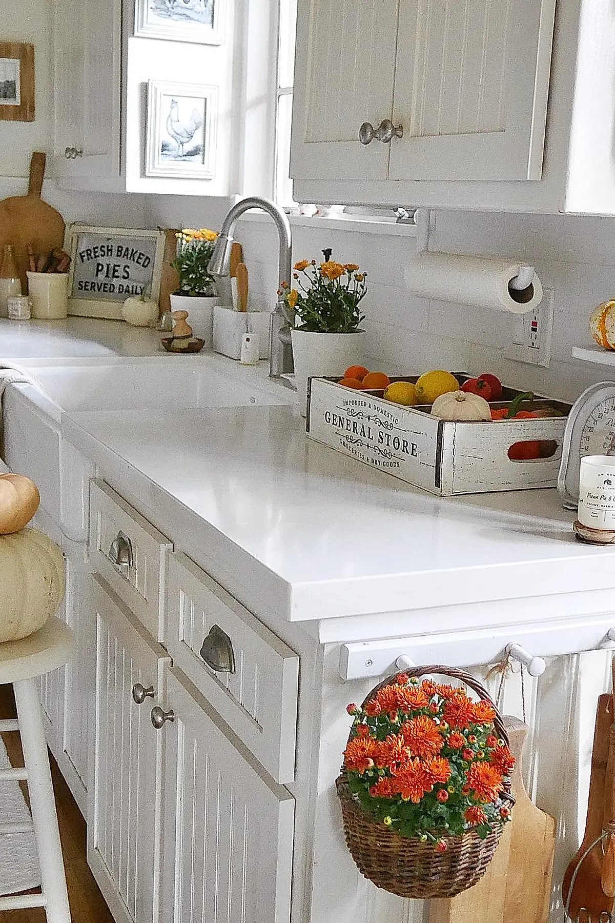White kitchen cabinets and countertop with a wicker basket of orange chrysanthemums hanging on the side, alongside a wooden crate filled with colorful fruit including oranges, lemons, and small pumpkins.