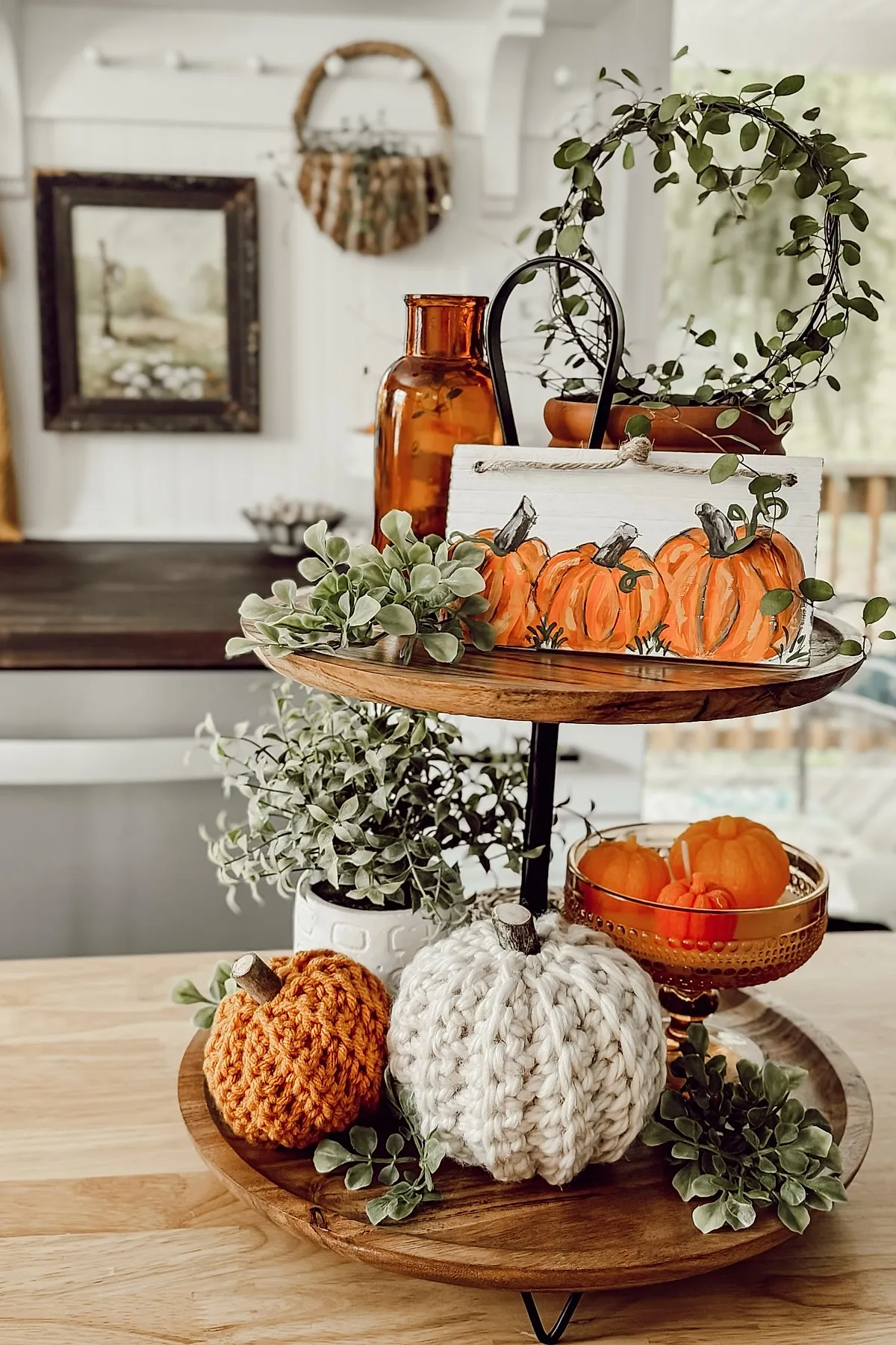 Two-tier wooden stand displaying cream and orange knit pumpkins, amber glass bottle and bowl with small orange pumpkins, leafy greenery sprigs, and a wreath wrapped around the top handle in a bright kitchen.