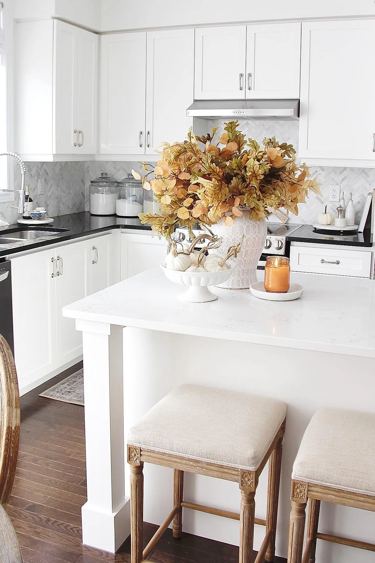 White kitchen island topped with an oversized white ceramic vase holding golden and muted orange fall foliage, alongside a pedestal bowl of small white pumpkins and an amber candle in a glass jar.