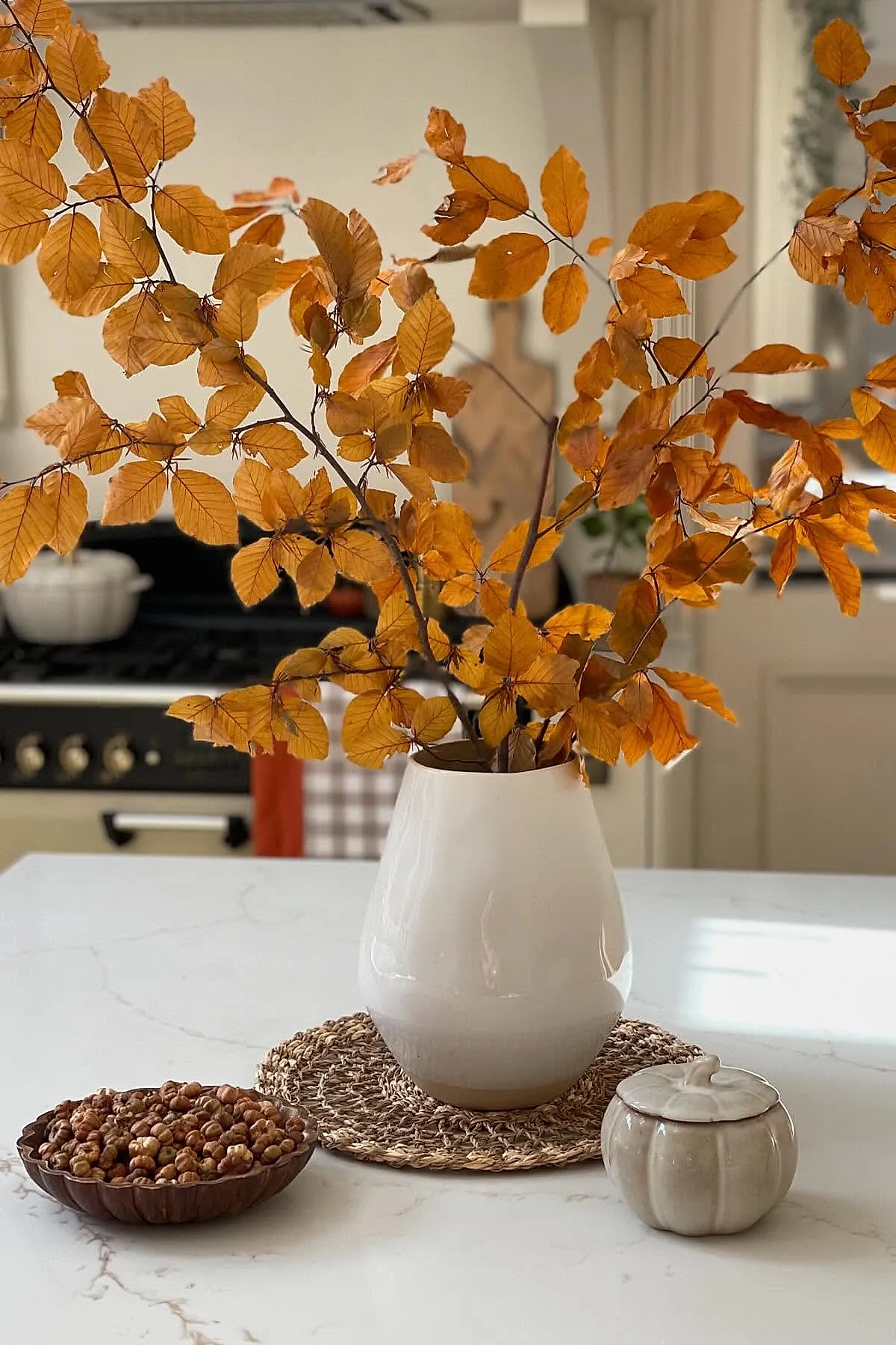 White ceramic vase holding large branches with bright orange fall leaves on a white kitchen island, next to a woven placemat, a small ceramic pumpkin container, and a wooden bowl filled with nuts.
