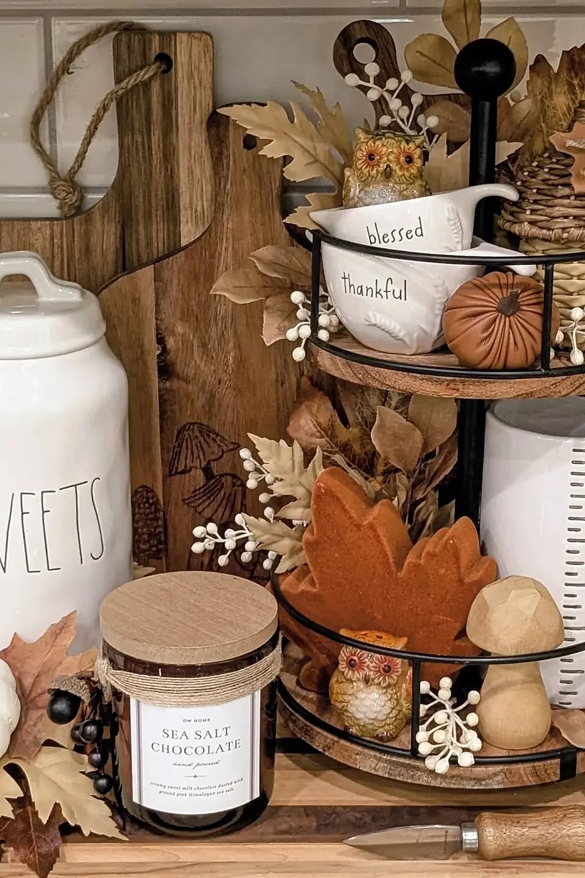 Two-tier wooden and metal tray holding white ceramic bowls labeled blessed and thankful, surrounded by faux beige autumn leaves, small leather pumpkin, wicker pinecone, miniature owl figurines, and a wooden mushroom on a kitchen countertop.