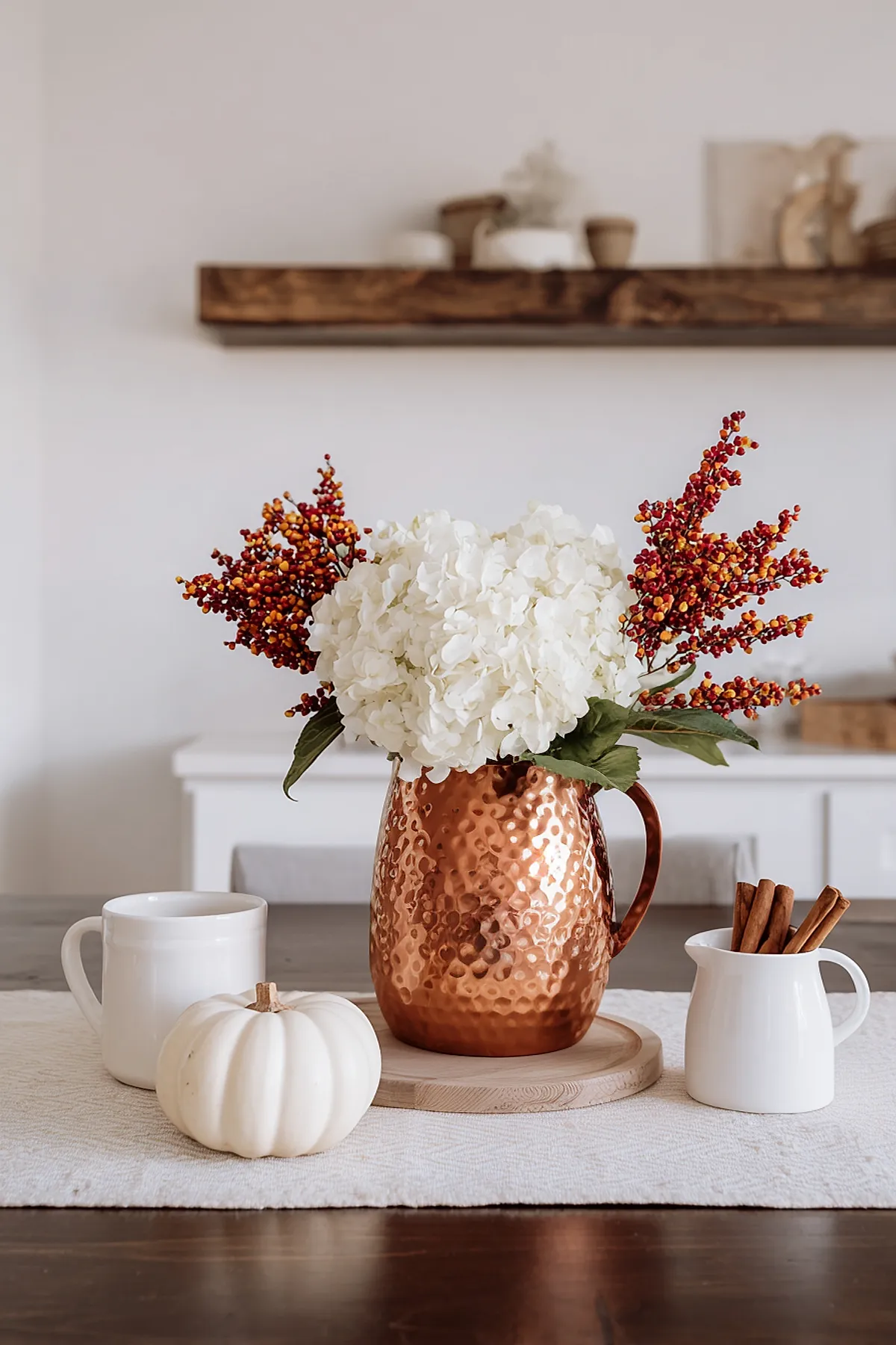 Hammered copper pitcher filled with white hydrangeas and red berry branches on a light kitchen table alongside a small white pumpkin, a white mug, and a small ceramic jug holding cinnamon sticks.