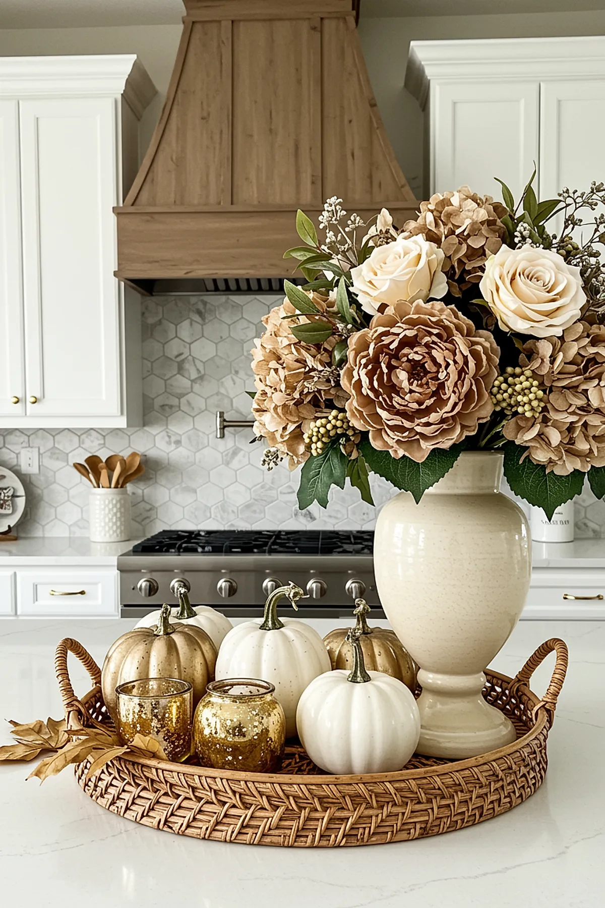 Woven round tray holding white and gold decorative pumpkins alongside a large beige ceramic vase filled with cream roses, tan hydrangeas, greenery, and small berries on a white marble kitchen island with wooden range hood in the background.