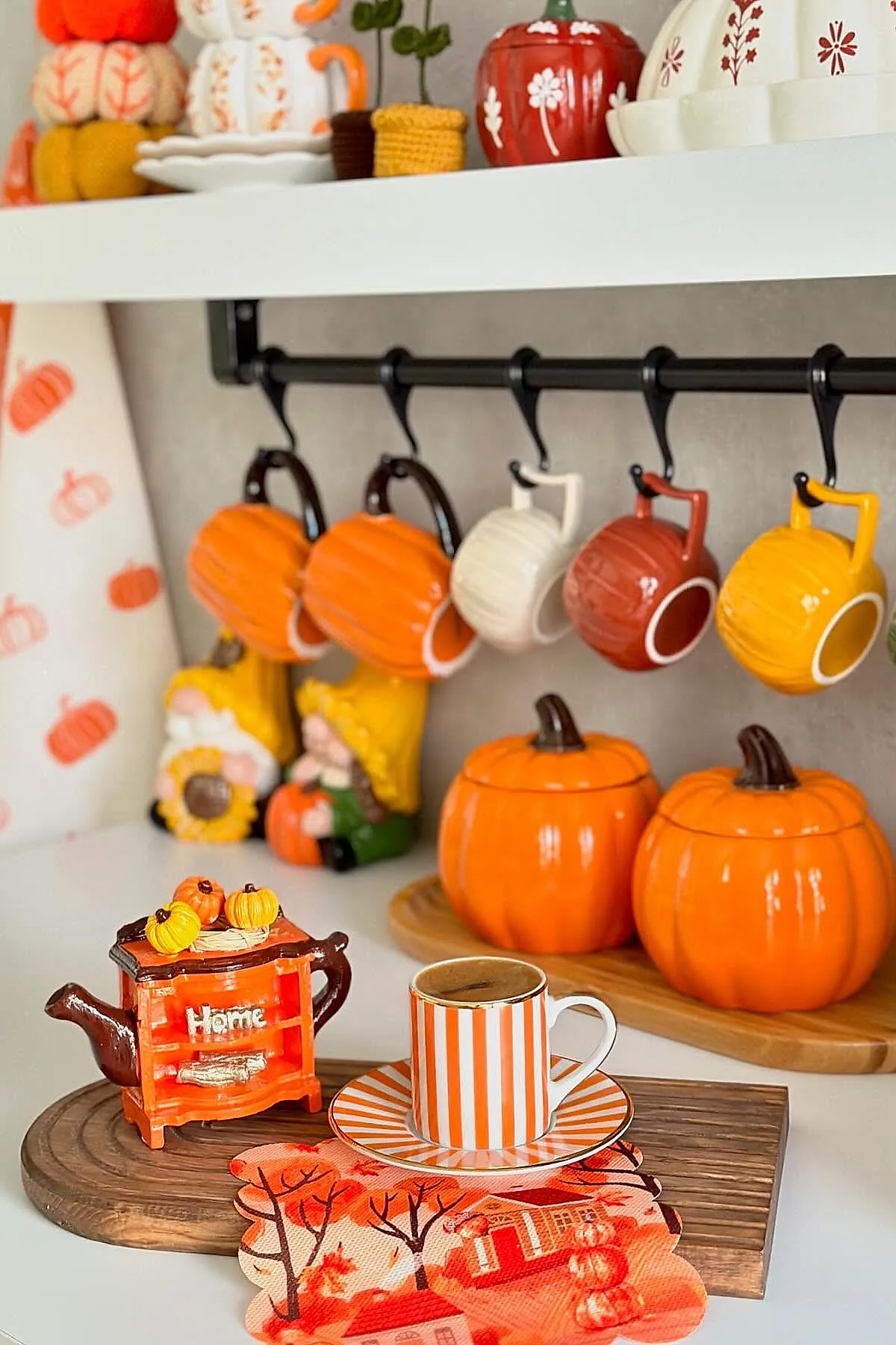 Kitchen countertop featuring orange, yellow, red, and white pumpkin-shaped mugs hanging on black hooks, two ceramic pumpkin containers on a wooden board, an orange and white striped espresso cup on saucer, and a small decorative teapot with tiny pumpkins.