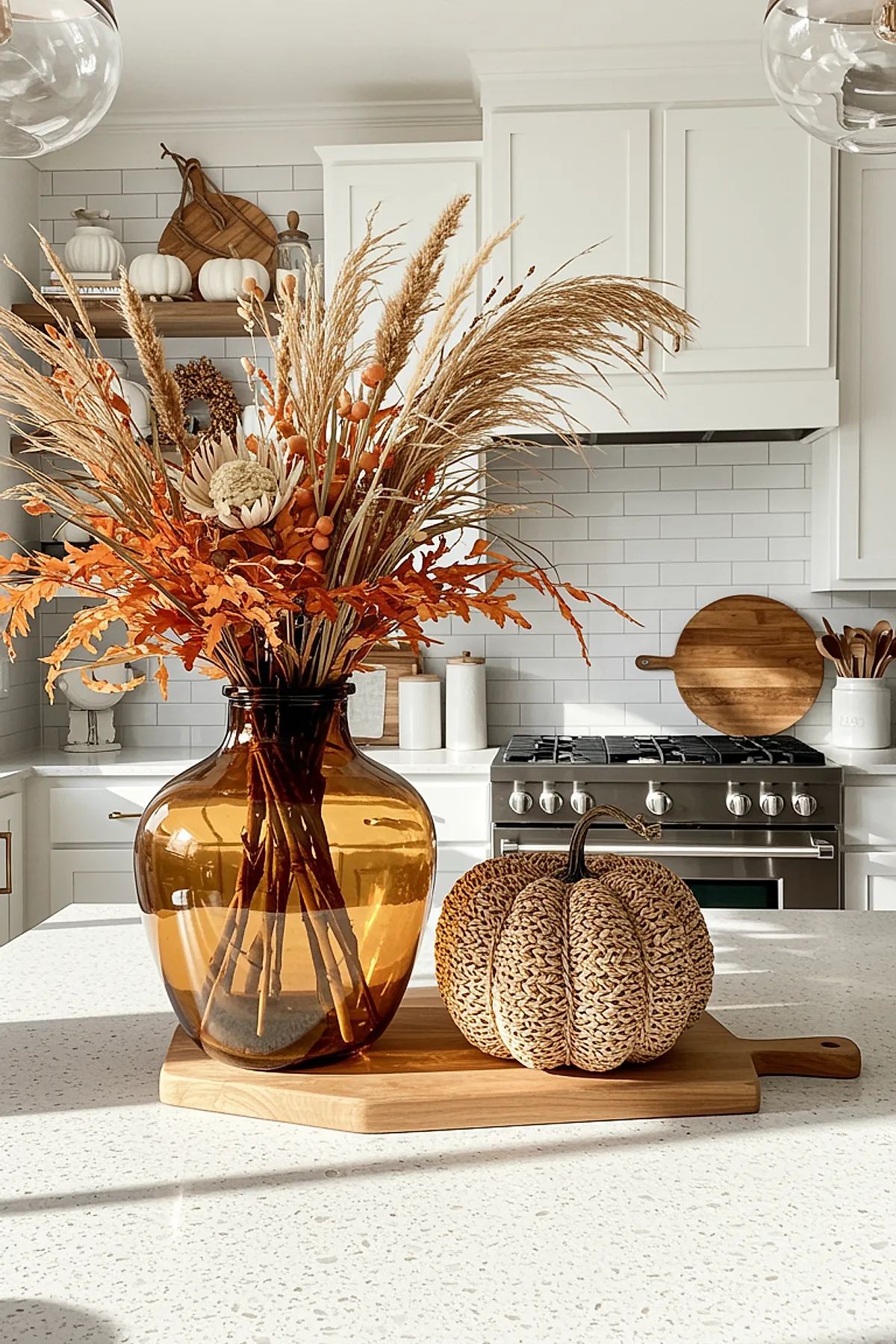 Amber glass vase filled with dried pampas grass, orange leaves, and seed pods next to a woven rattan pumpkin placed on a light wooden cutting board on a white kitchen island.