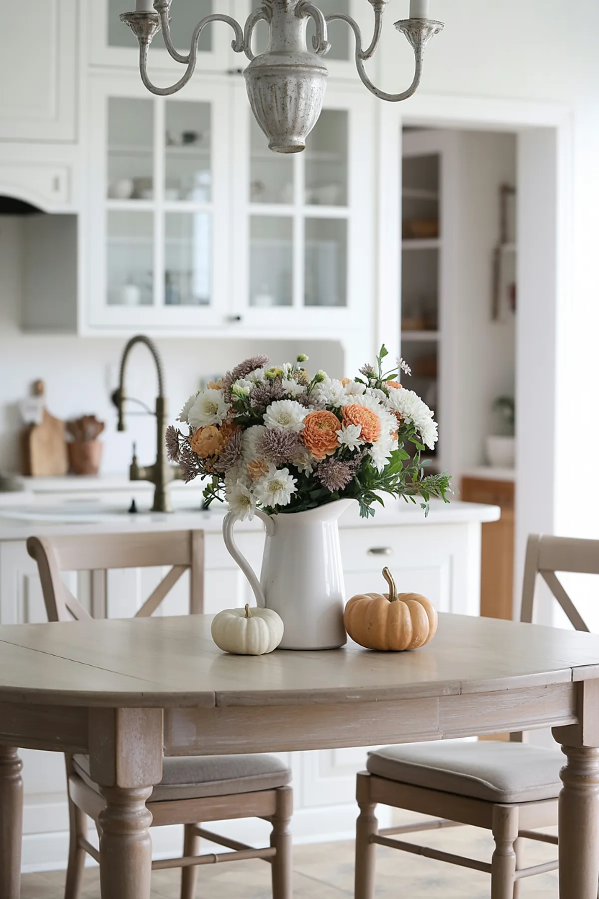 Light wood kitchen table with a white ceramic pitcher holding mixed cream, orange, and lavender flowers, flanked by two small pumpkins-one white and one pale orange-against a bright kitchen background.