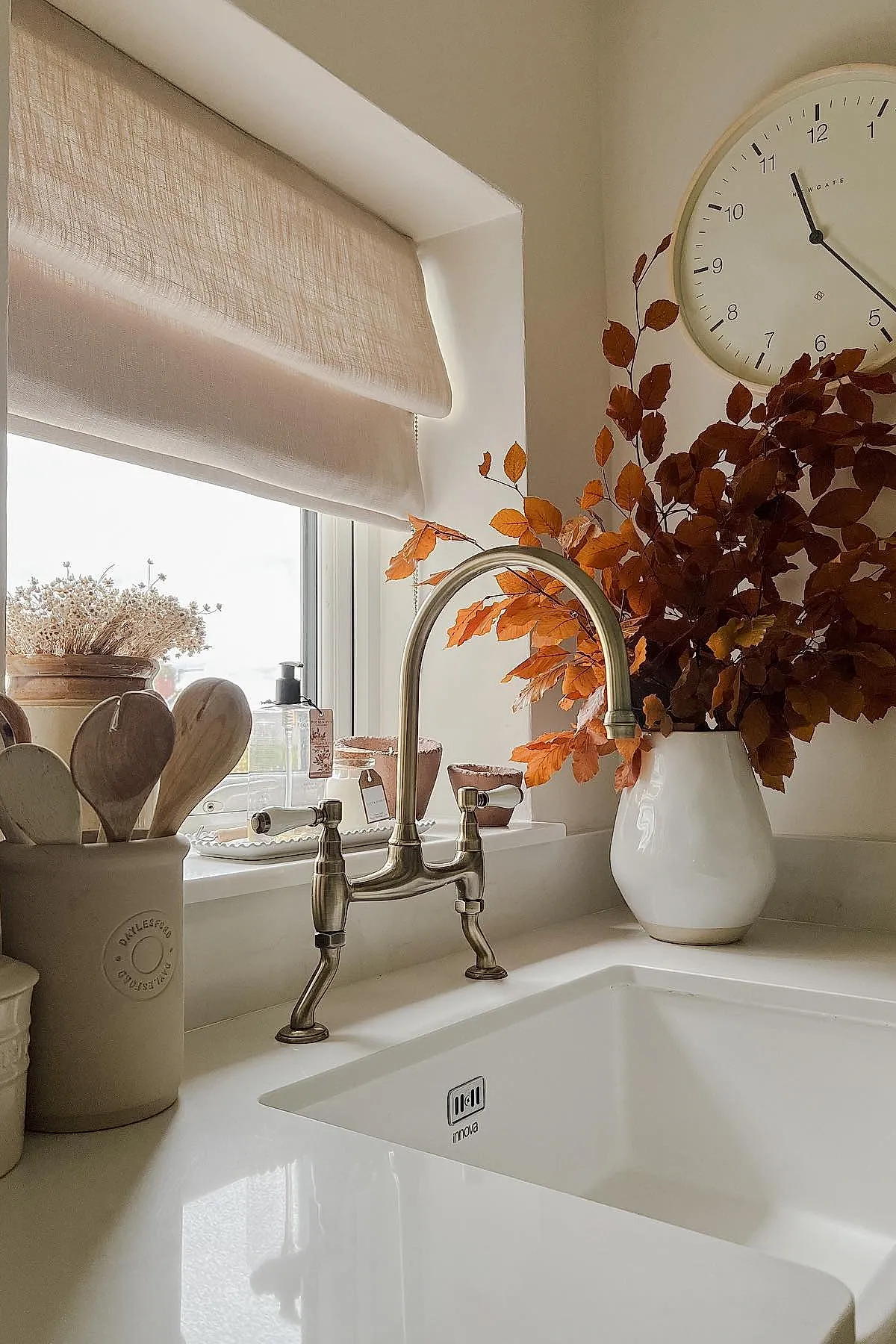 White ceramic vase holding vibrant orange autumn leaves positioned next to a kitchen sink under a beige roman shade, with wooden cooking utensils in a ceramic holder on the left side.