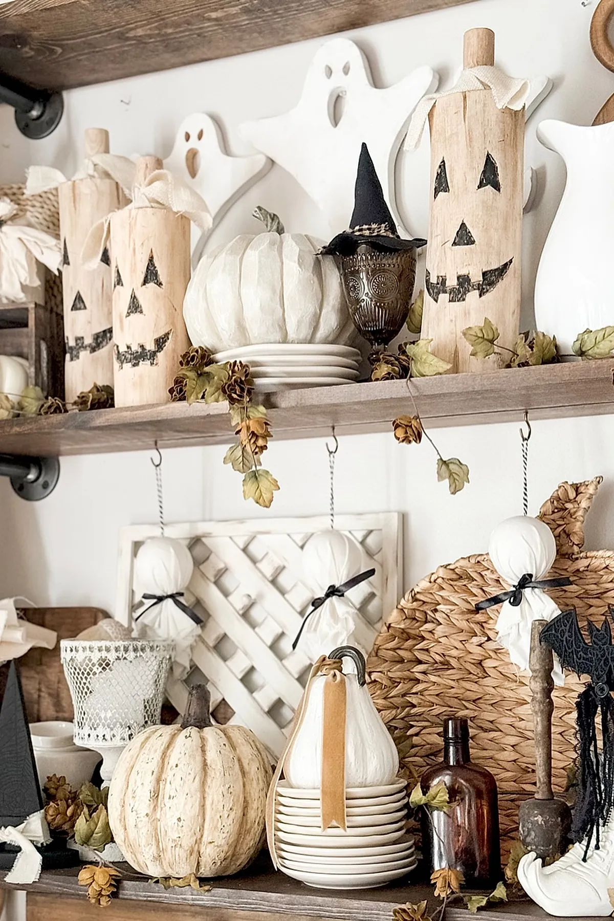 Open rustic wooden kitchen shelves displaying carved wooden jack o lantern blocks, white ghost cutouts, white pumpkins, pine cones, dried leaves, and vintage glassware.