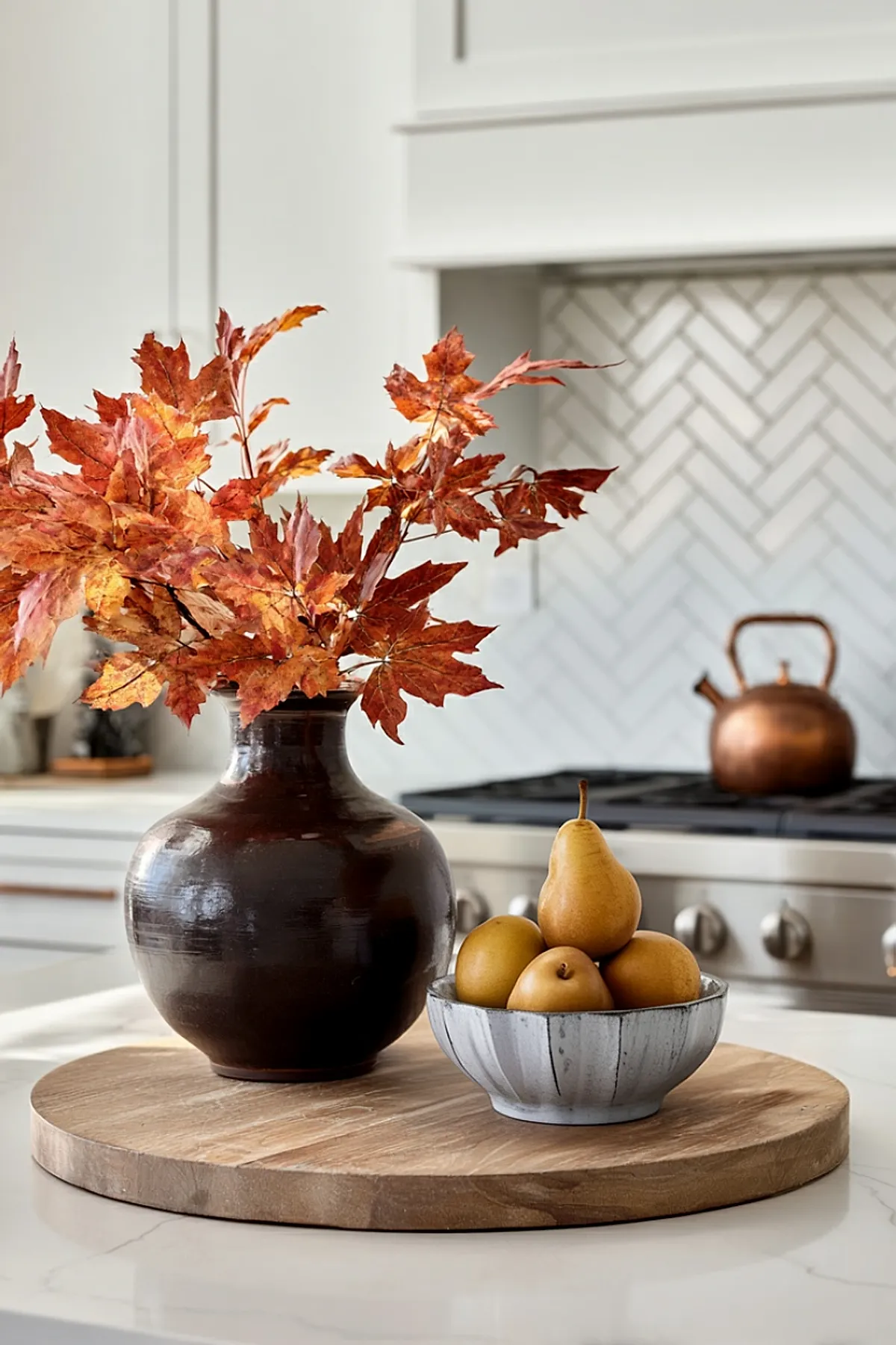 Dark brown ceramic vase holding bright orange fall maple leaves next to a grayish white bowl filled with yellow Asian pears, all placed on a round wooden tray on a light-colored kitchen countertop.