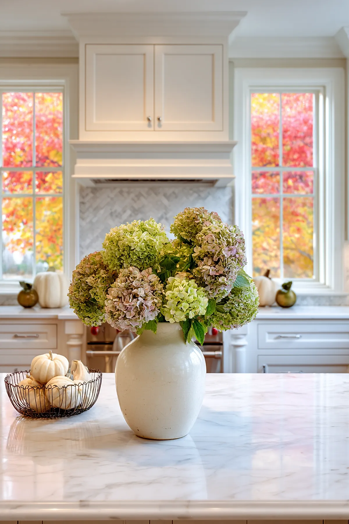 White marble kitchen island topped with a large cream ceramic vase filled with green and pale pink hydrangeas, next to a wire basket holding small white pumpkins. Bright fall foliage in red, orange, and yellow is visible through two windows in the background.
