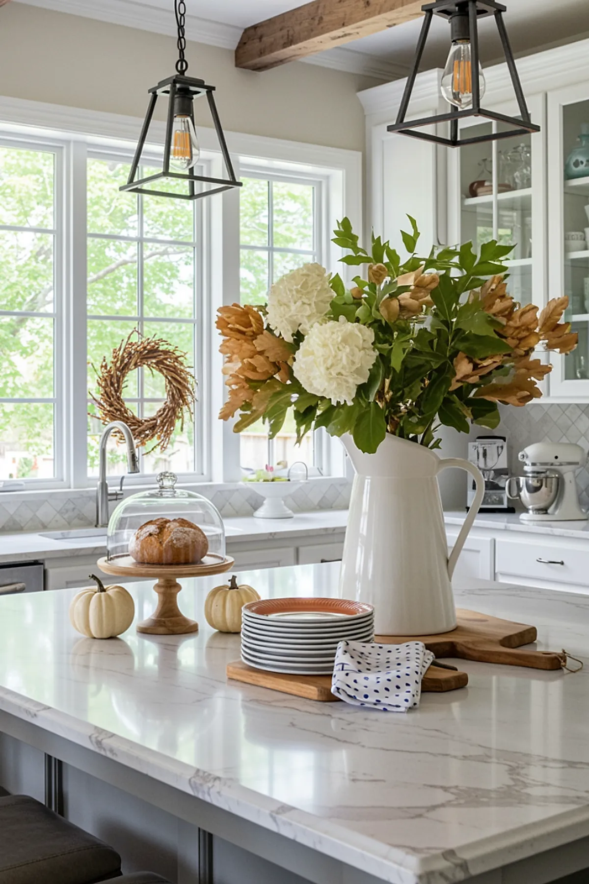 Bright kitchen island featuring a large white ceramic pitcher holding green leaves, white hydrangeas, and brown dried foliage, flanked by two small white pumpkins on a marble countertop near large windows.