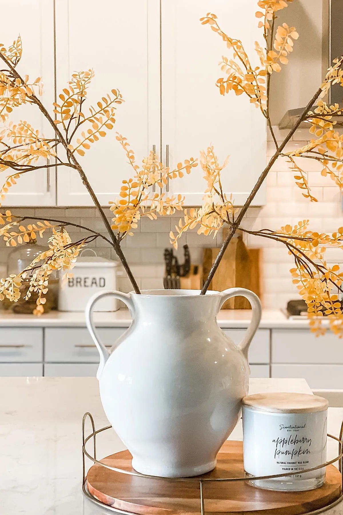 White two-handled ceramic vase holding thin branches with small orange leaves, sitting on a round wooden tray next to an appleberry pumpkin scented candle in a bright kitchen.
