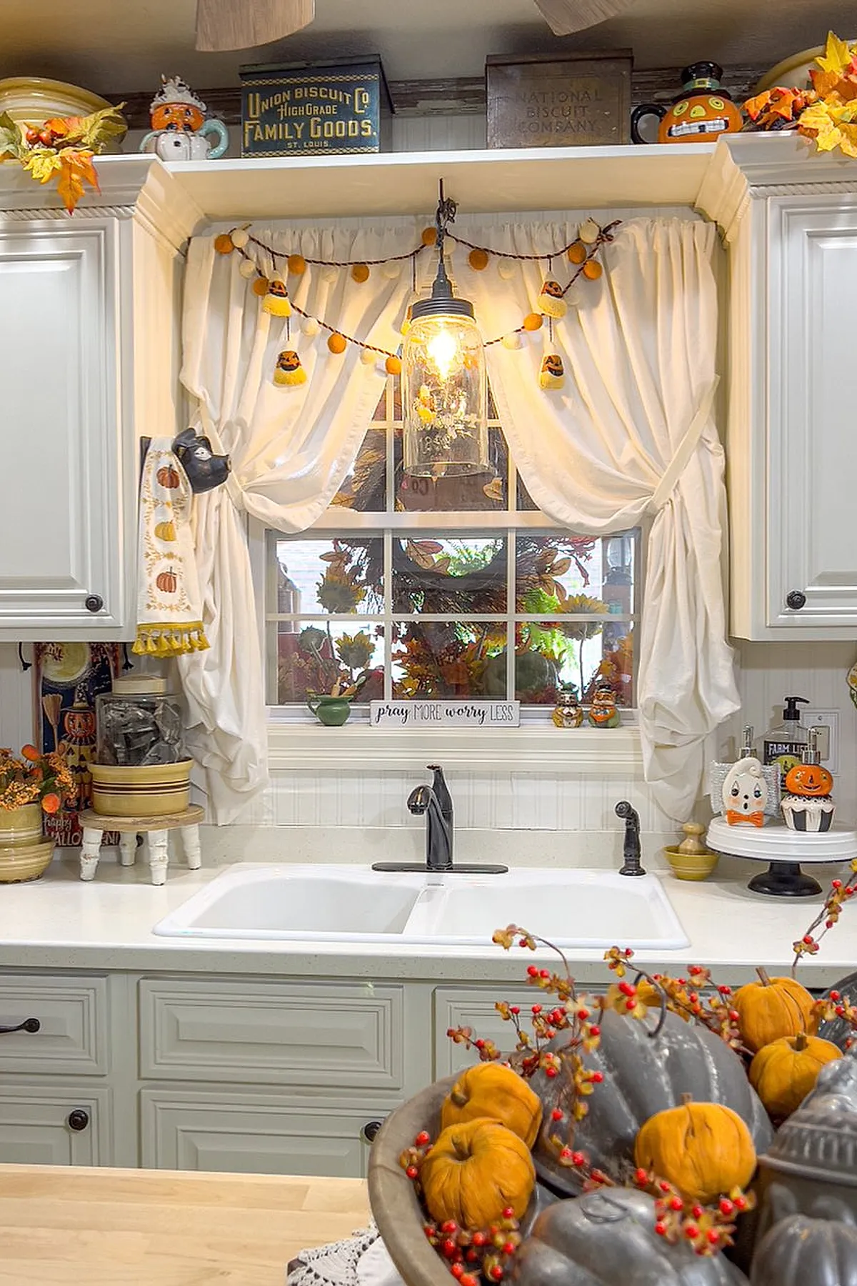 Kitchen sink area with white cabinetry, creamy gathered curtains tied back, orange felt pumpkin garland hanging across the window, autumn wreath visible outside, small fall figurines and a wooden sign on the windowsill.