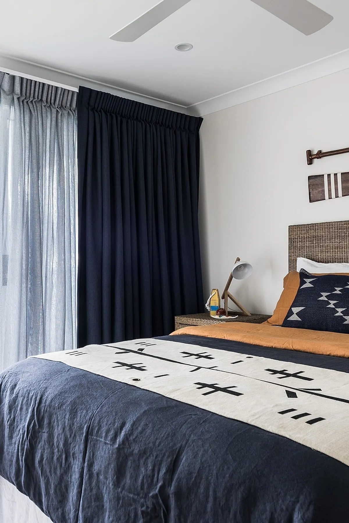 Teen boy bedroom featuring a bed with layered bedding including navy, white tribal pattern blanket, burnt orange sheets, woven rattan headboard, dark navy curtains, white ceiling fan, and wooden paddle wall art.