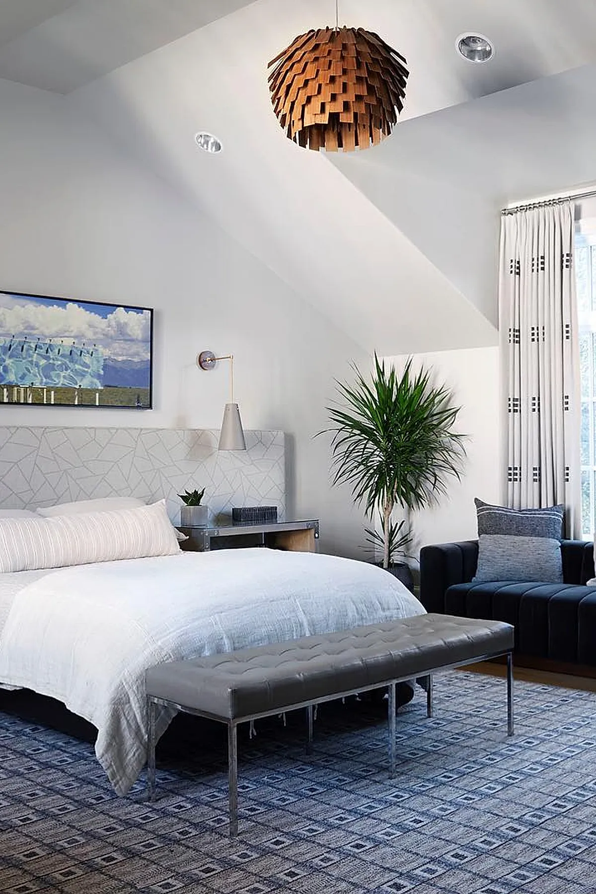 Teen boy bedroom with white walls, geometric upholstered headboard, gray tufted leather bench, blue patterned rug, black sofa with gray pillows, tall green plant, and a textured wooden pendant light hanging from ceiling.