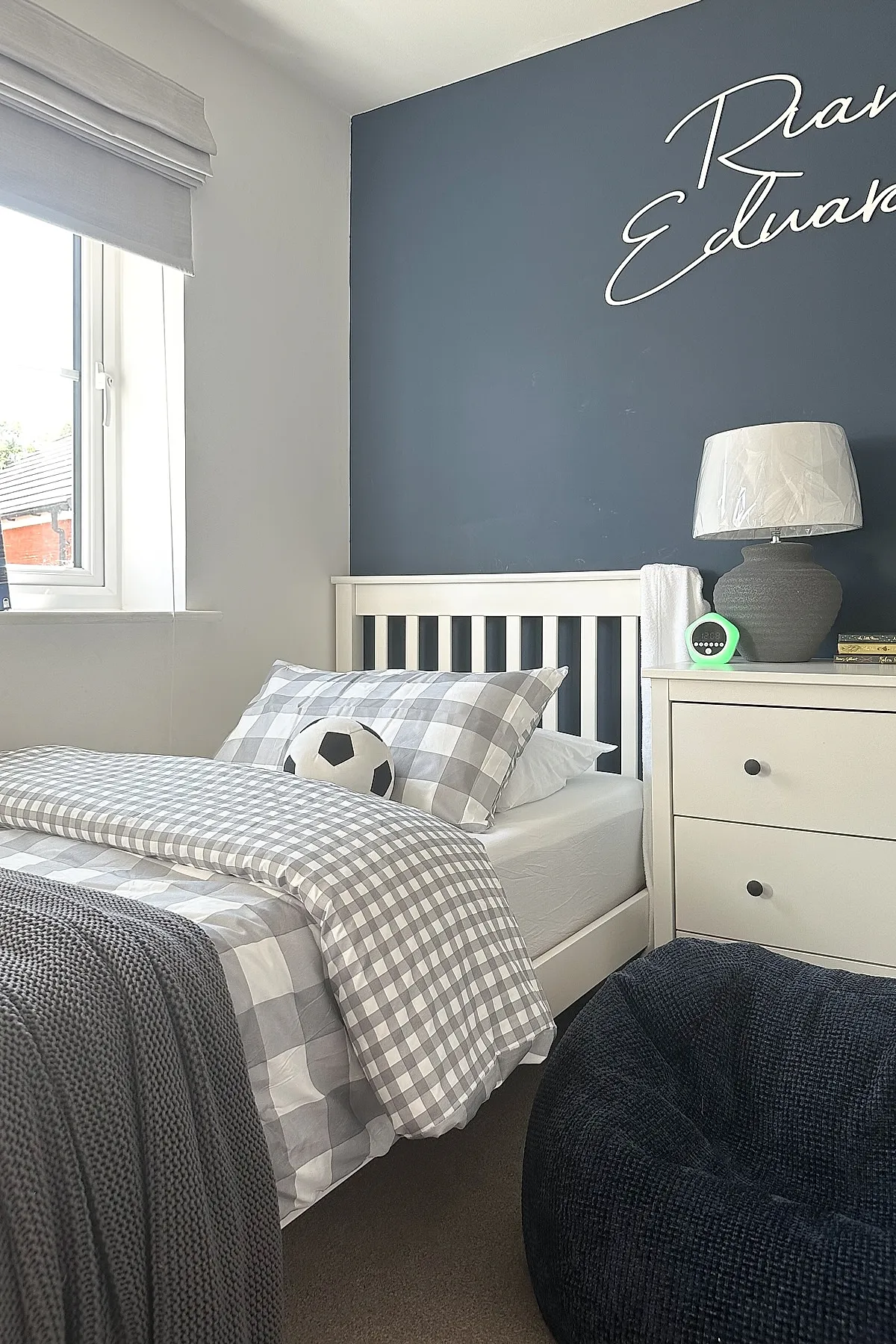 Teen boy bedroom featuring a navy blue accent wall, white bed frame with gray and white checkered bedding, a dark knit throw blanket, bean bag chair in dark textured fabric, and a bedside table with lamp.