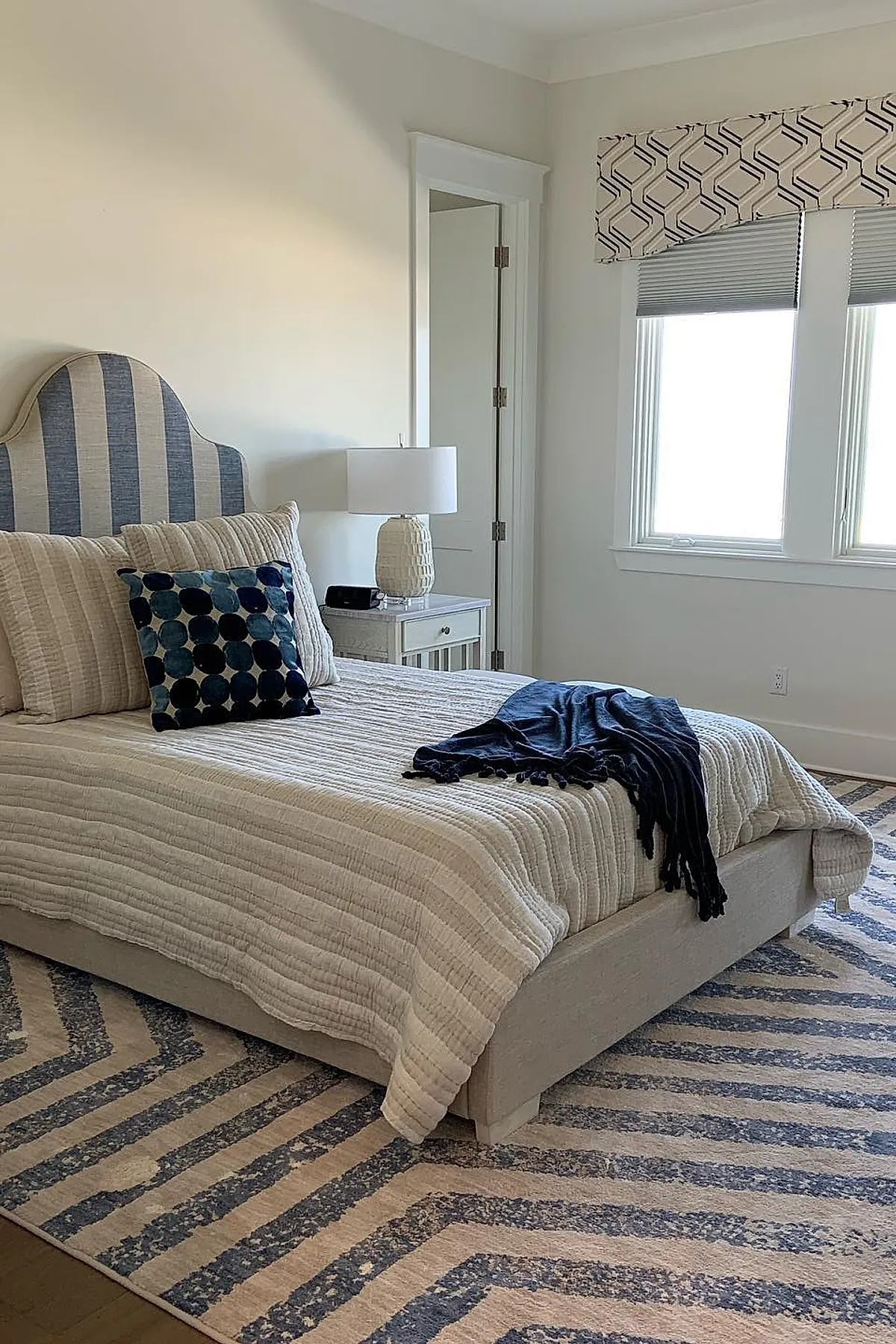 Teen boy bedroom featuring a bed with blue and beige vertical striped upholstered headboard, beige quilted bedding, navy blue throw blanket, polka dot pillow, white nightstand with textured white lamp, and windows with geometric patterned valance and blinds.