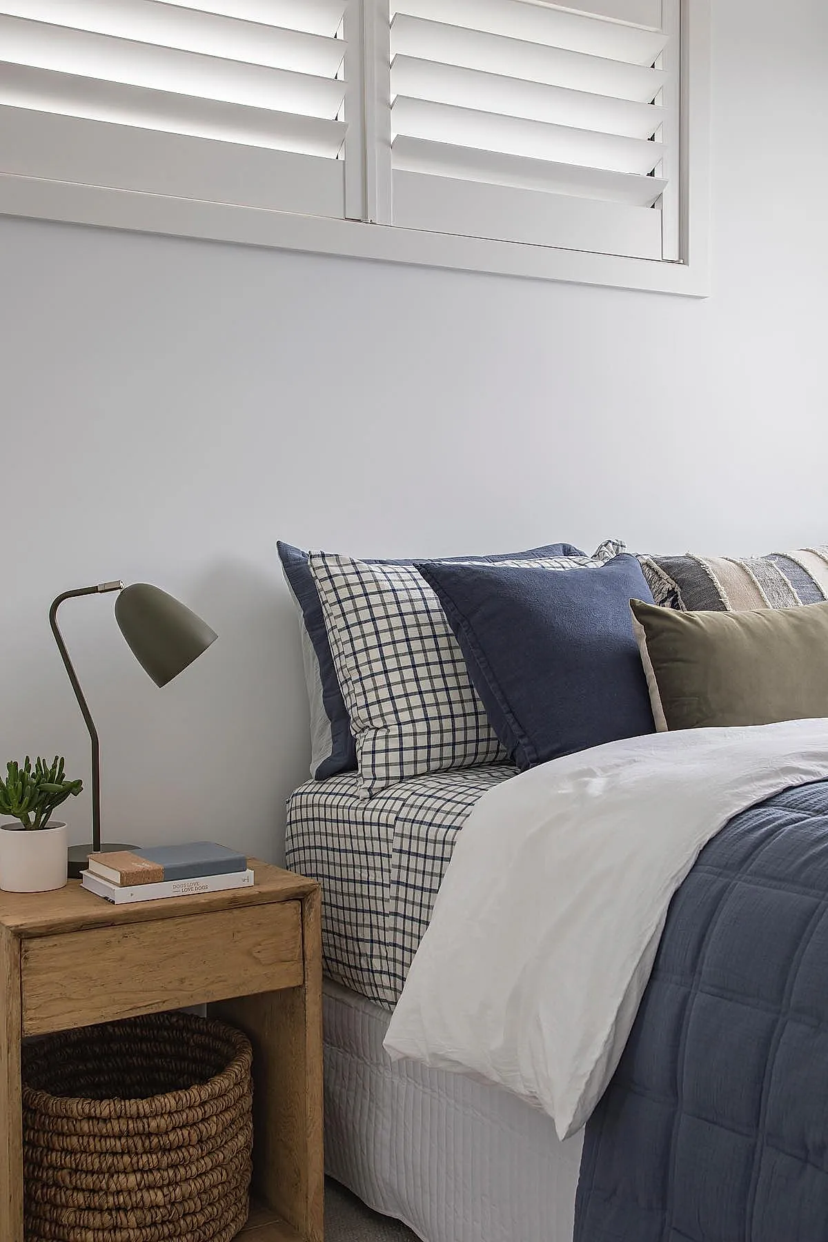Close-up of a bedroom corner featuring a wooden nightstand with an open shelf holding a round woven rattan basket, an olive green angled desk lamp, stacked books, and part of a bed with blue and white checkered bedding and layered pillows.