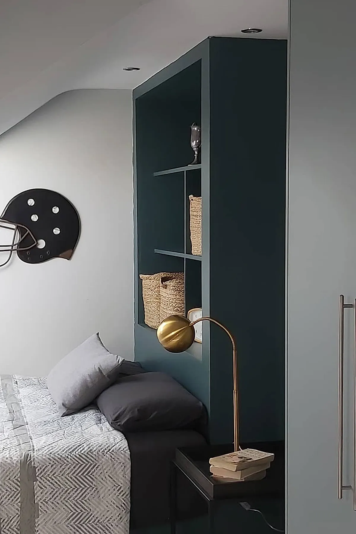Teen boy bedroom featuring a bed against deep teal built-in shelving headboard with woven baskets, gray bedding, brass gooseneck lamp on black nightstand, and football helmet wall decor.