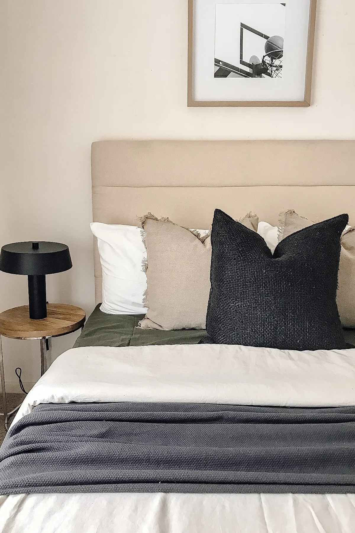 Bed with beige upholstered headboard, white sheets, layered beige and black textured pillows, next to a round wooden side table holding a matte black modern lamp, light beige walls with framed basketball hoop photo.