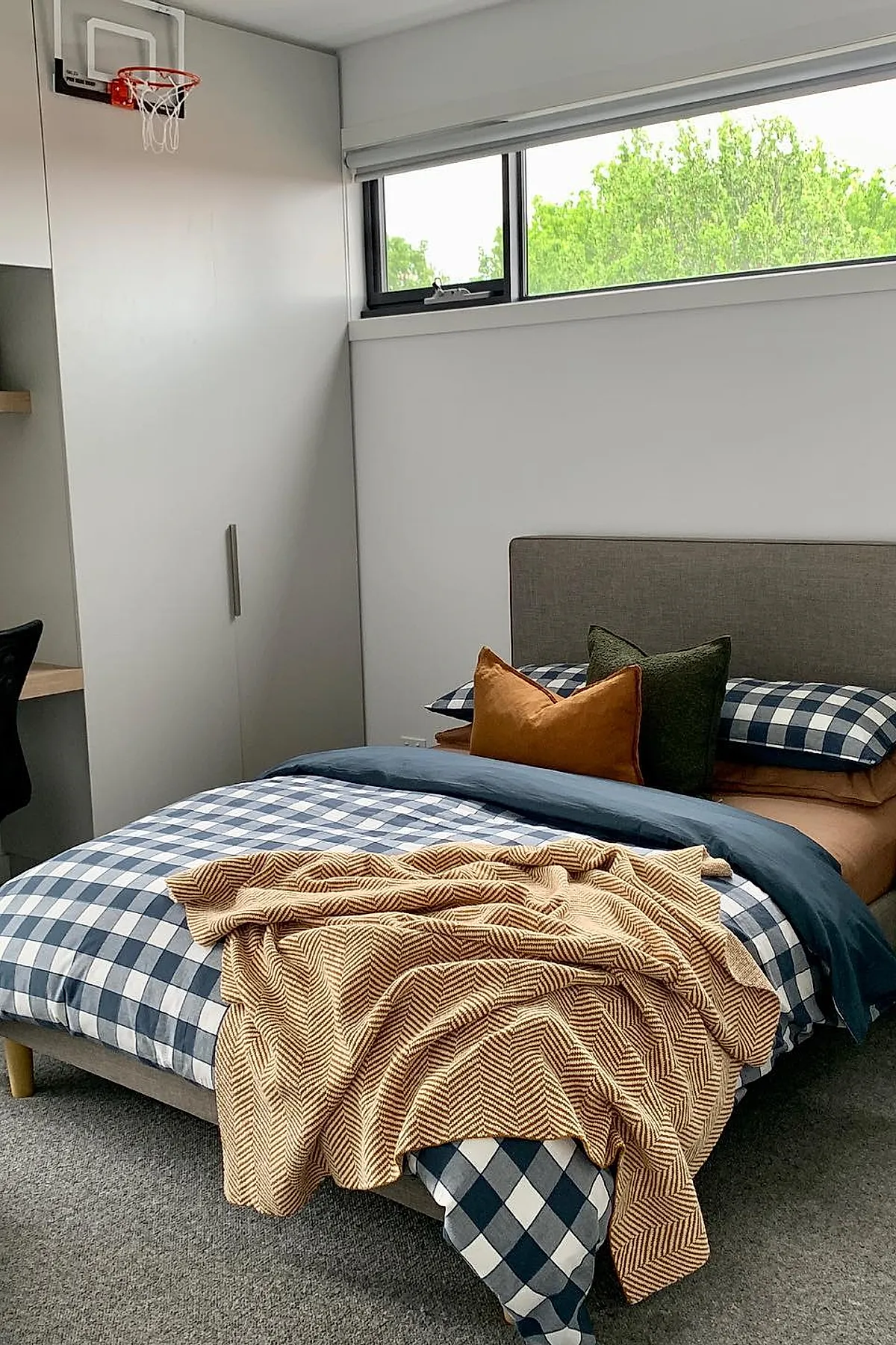 Teen boy bedroom featuring a bed with blue and white checkered bedding, orange and dark green pillows, brownish throw blanket, gray upholstered headboard, and a mini basketball hoop mounted on the wall above the closet door next to a high horizontal window showing greenery outside.